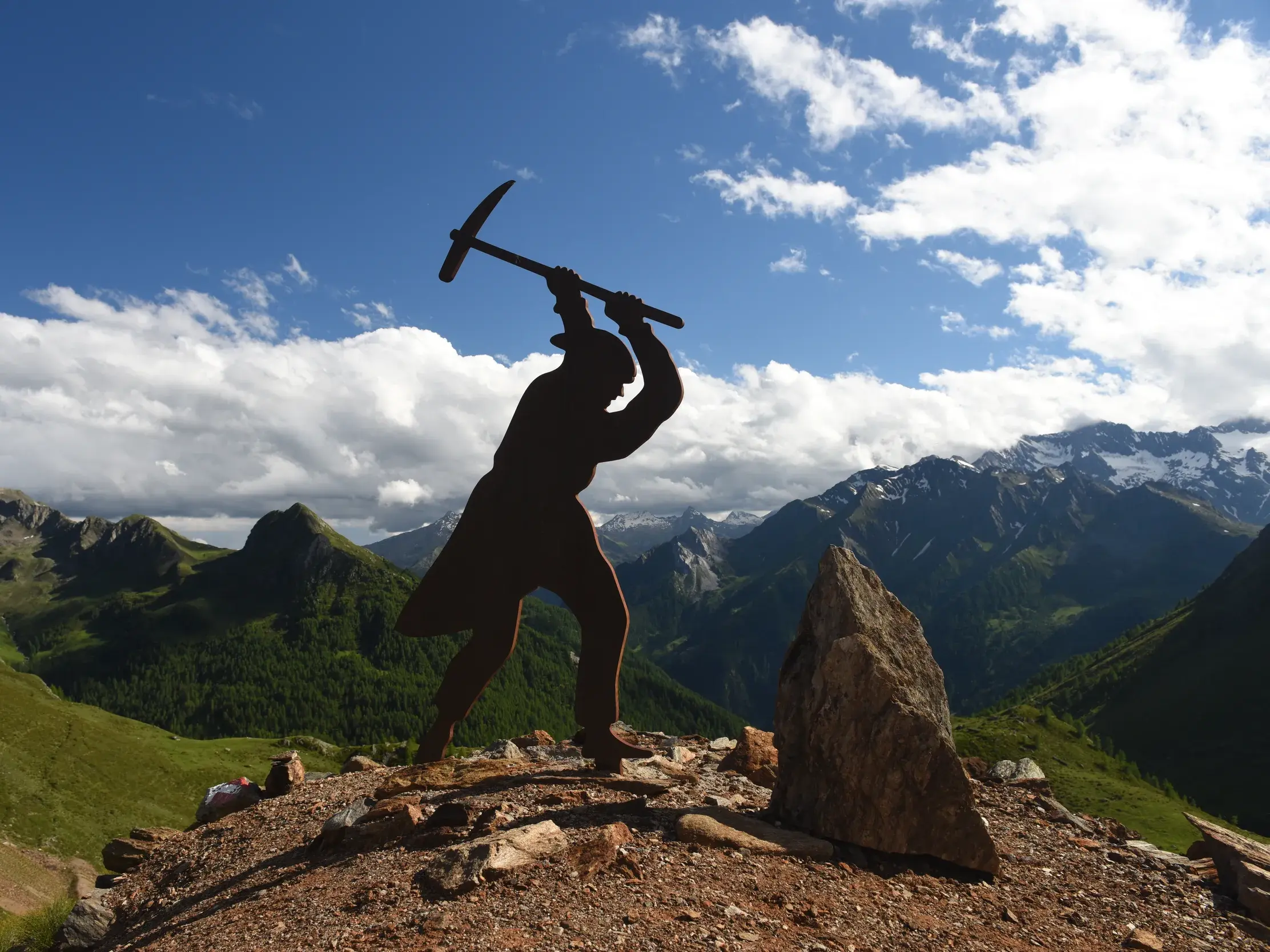 Silhouette einer Bergmann-Statue mit Spitzhacke vor einer beeindruckenden Berglandschaft mit grünen Tälern bei sonnigem Wetter.