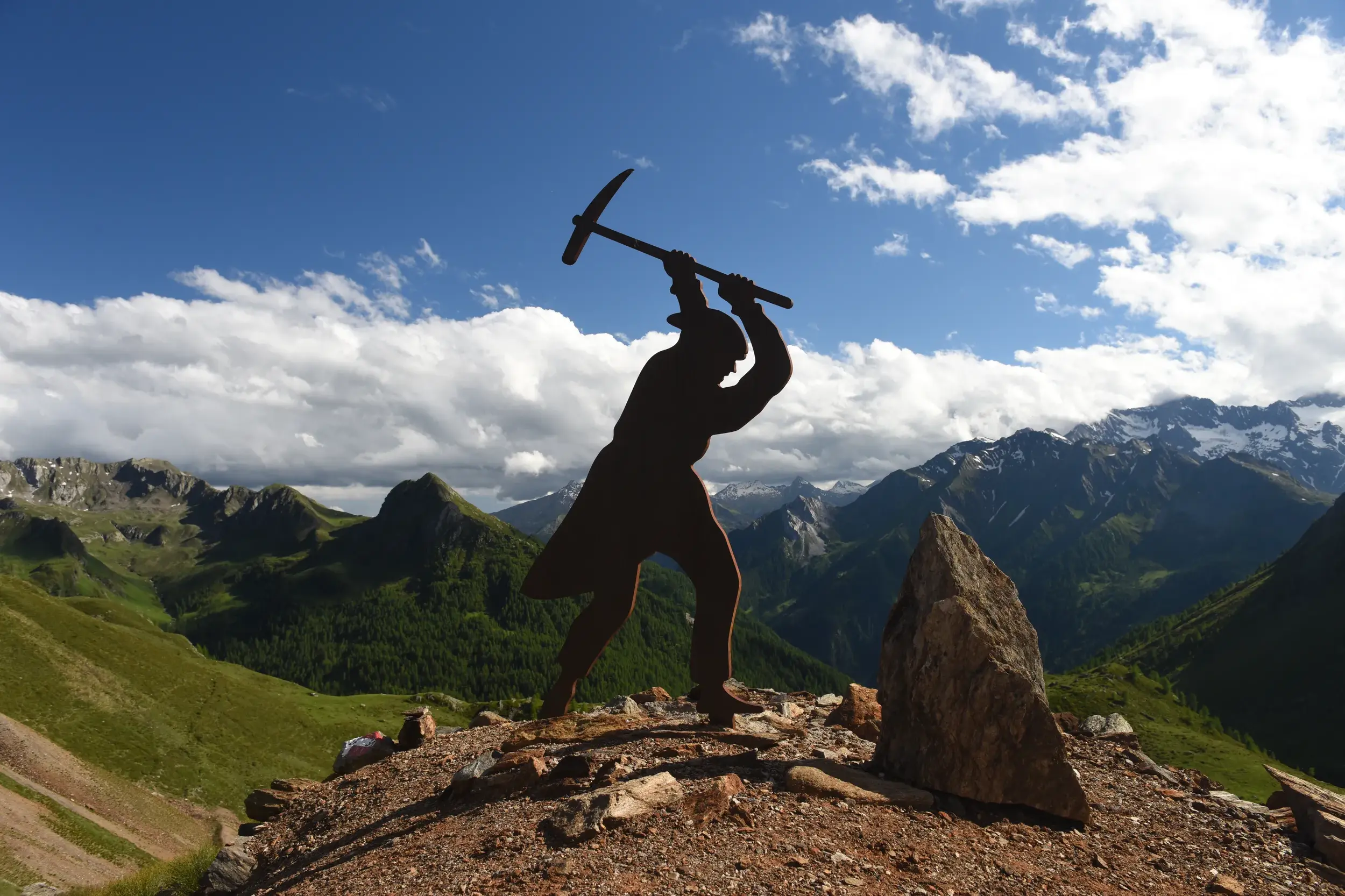 Silhouette einer Bergmann-Statue mit Spitzhacke vor einer beeindruckenden Berglandschaft mit grünen Tälern bei sonnigem Wetter.