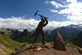Silhouette einer Bergmann-Statue mit Spitzhacke vor einer beeindruckenden Berglandschaft mit grünen Tälern bei sonnigem Wetter.