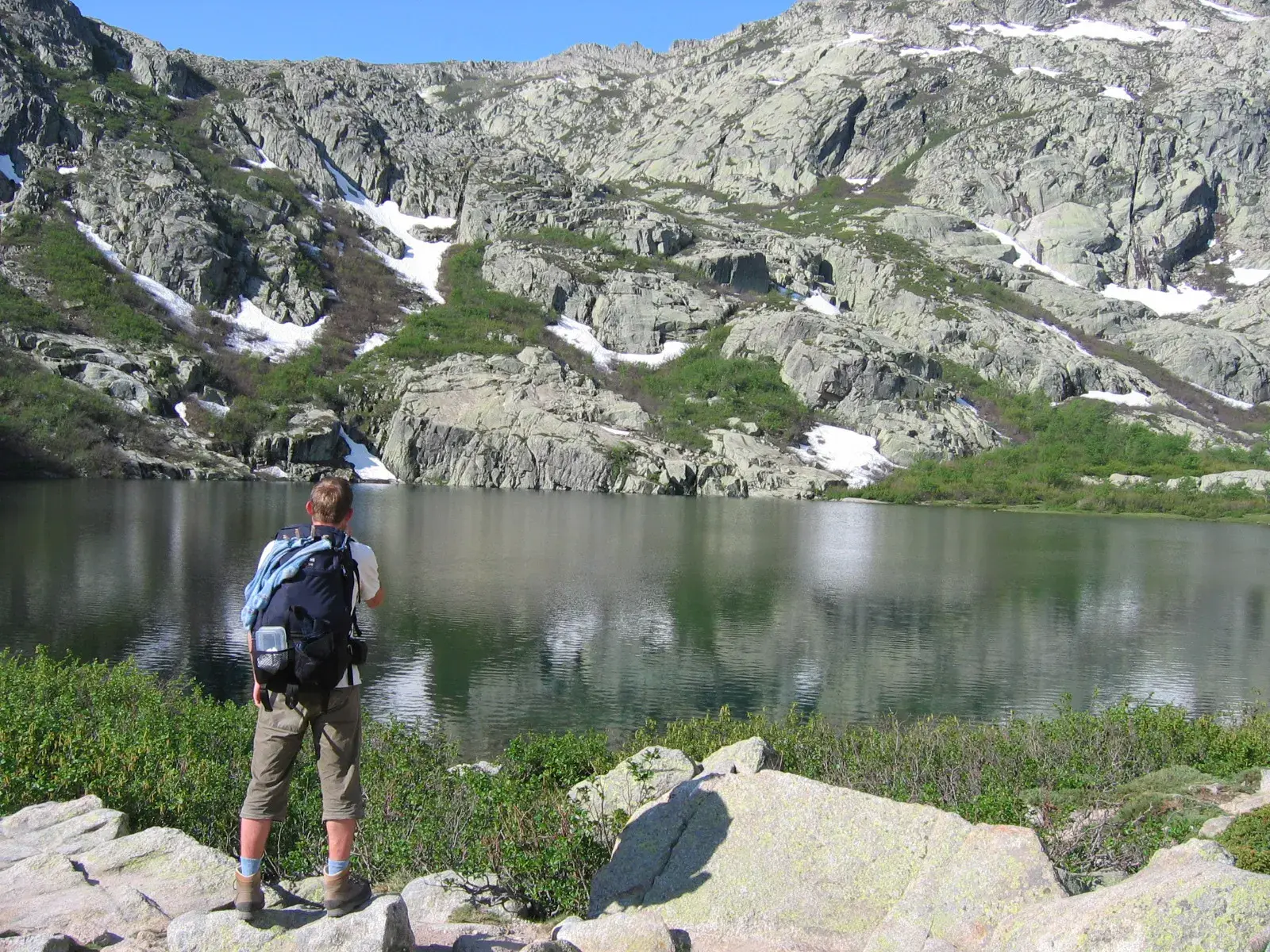 Wanderer mit Rucksack genießt die Aussicht auf einen Bergsee umgeben von schroffen Granitfelsen auf dem GR20 auf Korsika.