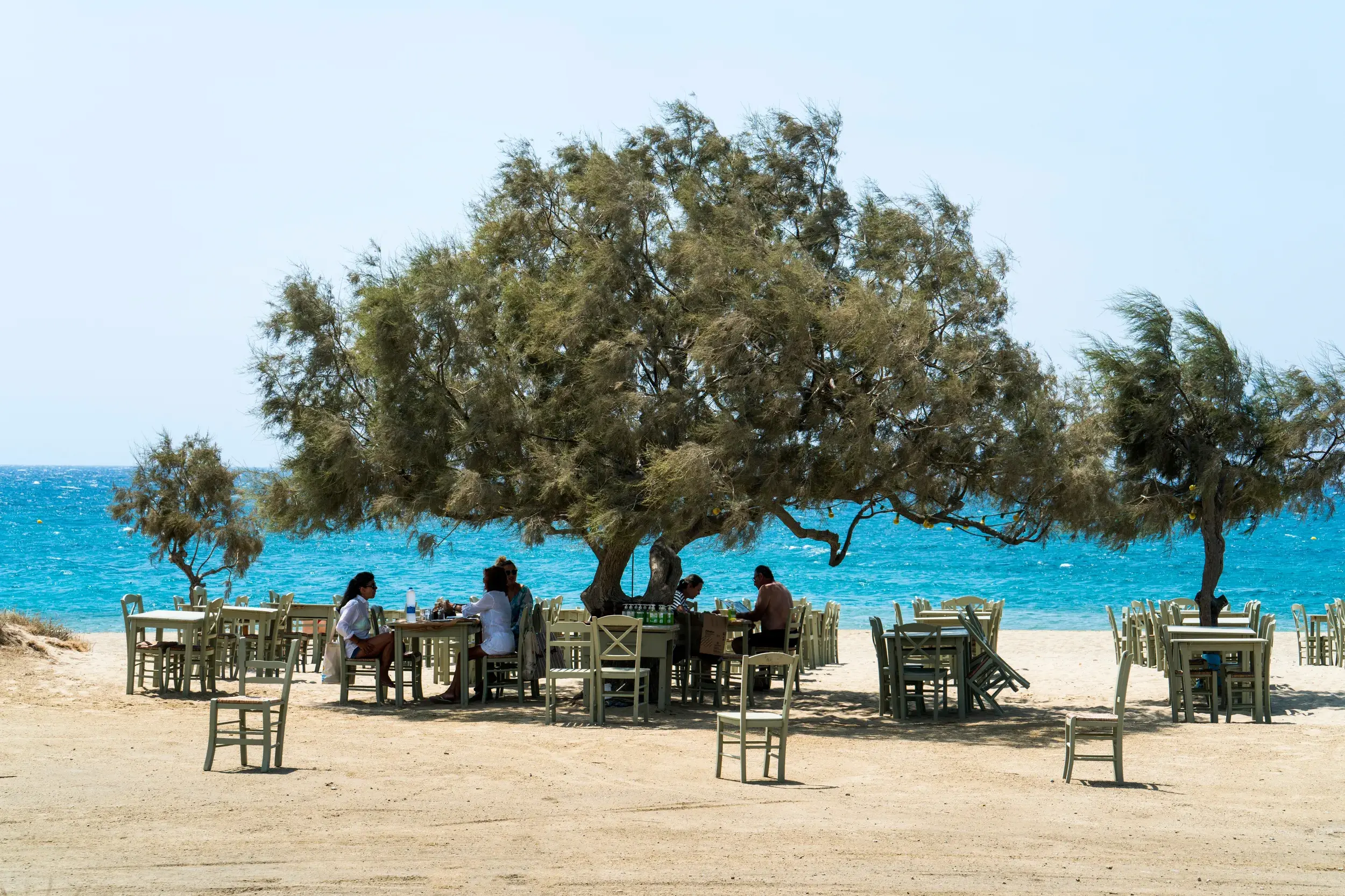 Eine kleine Gruppe genießt ihr Essen in einer Taverne am Sandstrand auf Naxos mit Blick auf das türkisblaue Meer.
