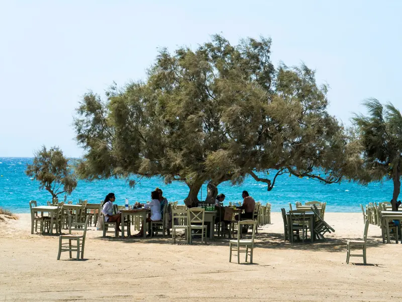 Eine kleine Gruppe genießt ihr Essen in einer Taverne am Sandstrand auf Naxos mit Blick auf das türkisblaue Meer.