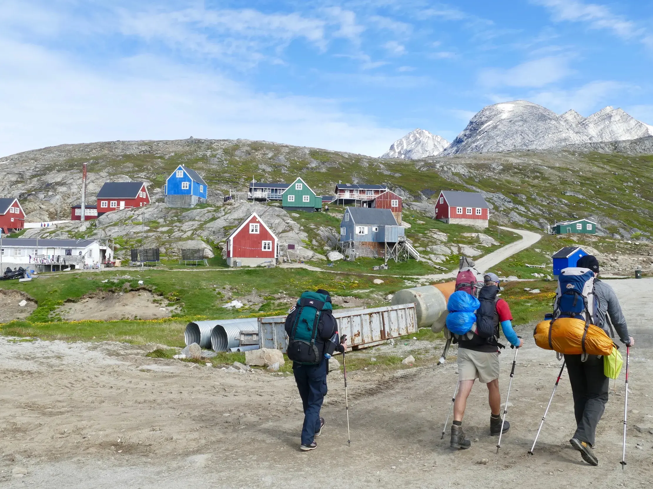 Eine kleine Trekking-Gruppe wandert mit Rucksäcken auf einem Schotterweg an bunten Häusern der Siedlung Tasiilaq in Grönland vorbei.