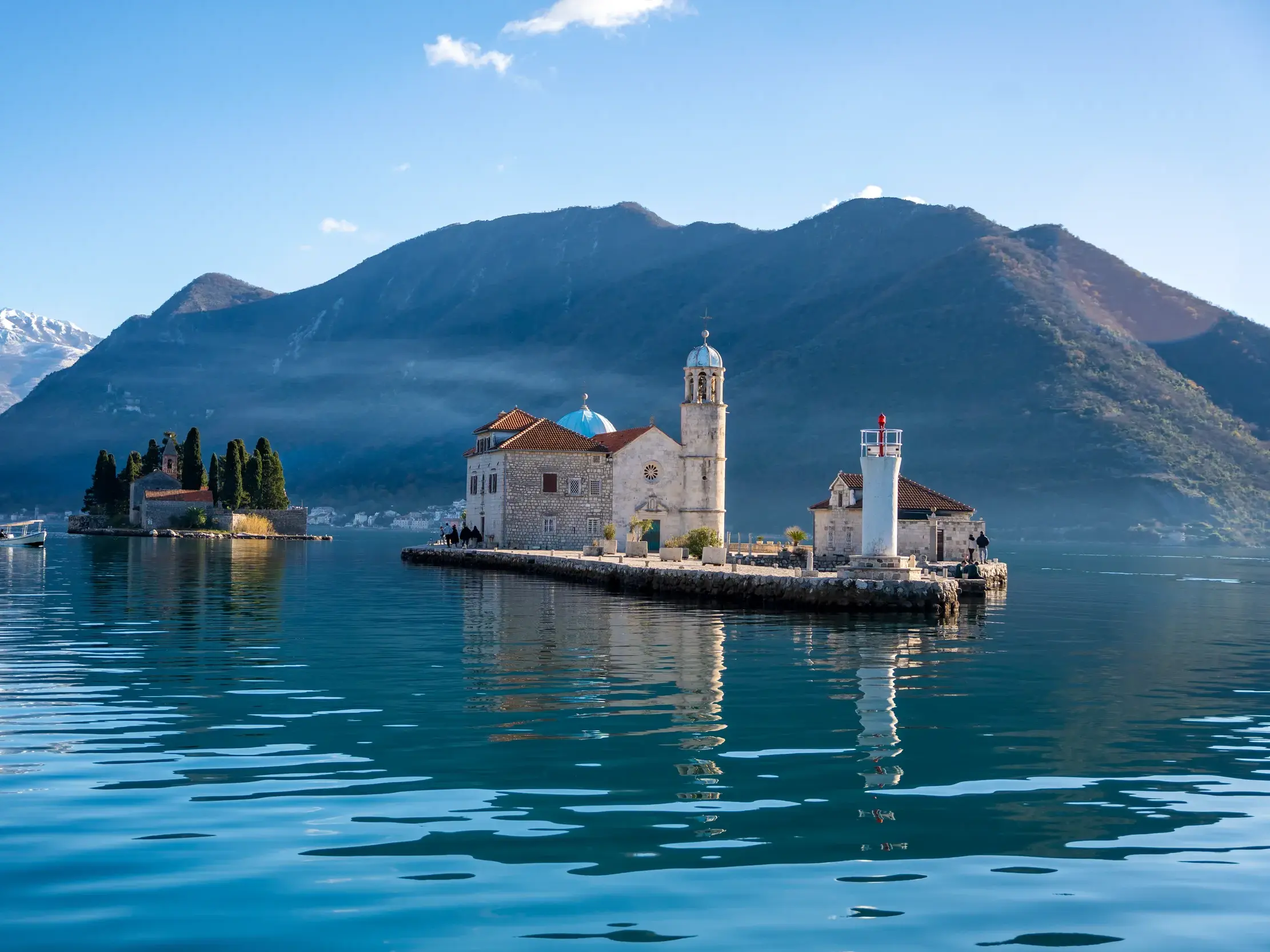 Bootsfahrt zur Insel Maria vom Felsen bei Perast mit Votivkirche in der Bucht von Kotor vor malerischer Bergkulisse.
