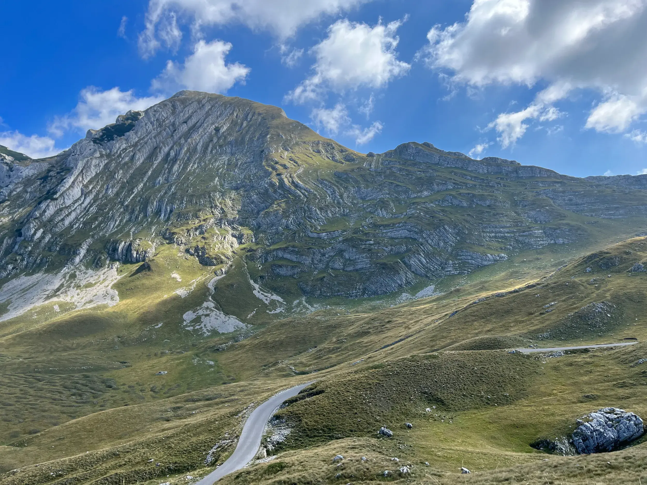 Eine schmale Straße führt durch die grünen Hügel zum schroffen Karstgipfel des Durmitor Nationalparks in Montenegro.