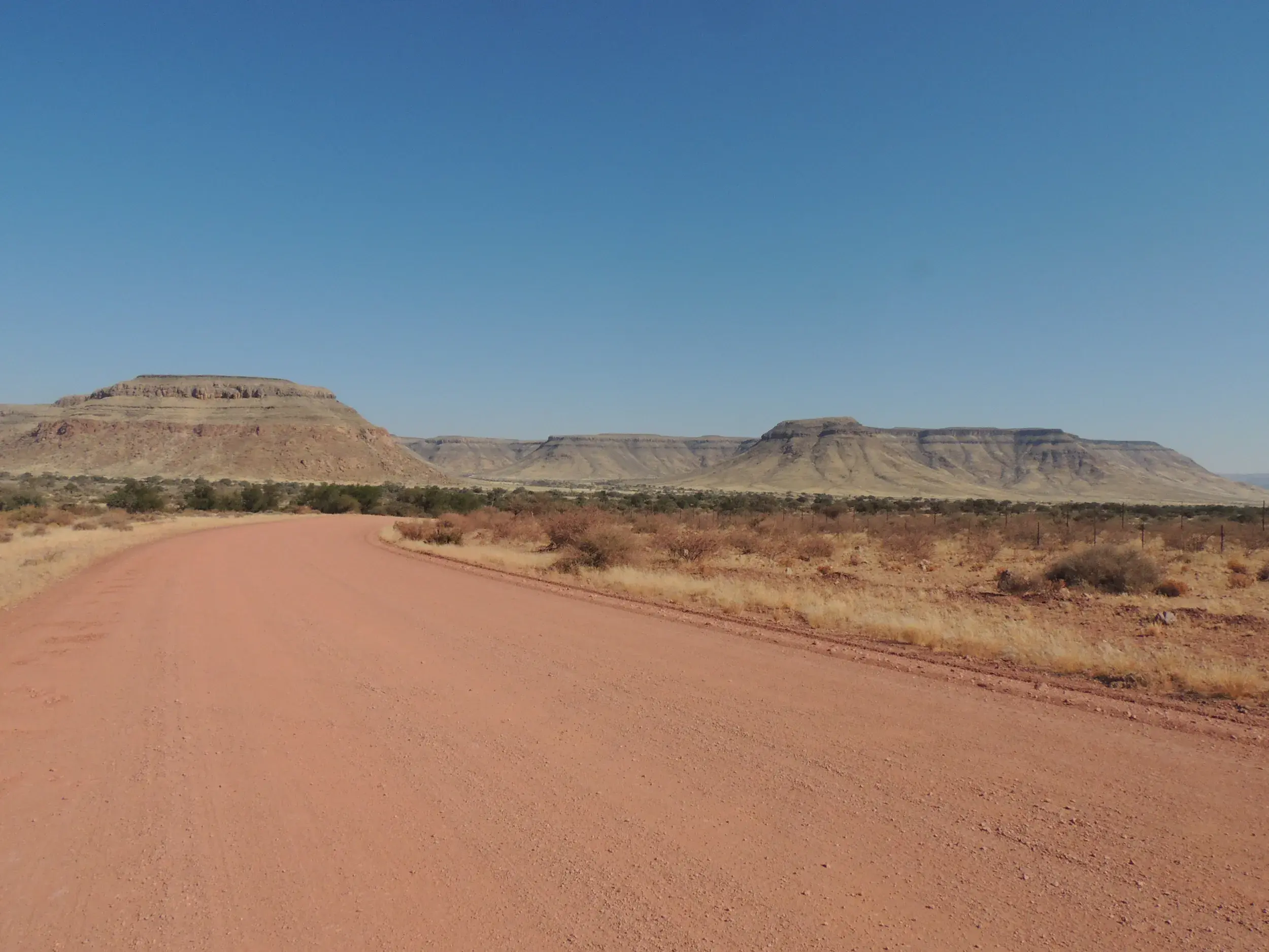 Eine rote Sandpiste führt durch die trockene Wüstenlandschaft Namibias mit markanten Tafelbergen am Horizont.