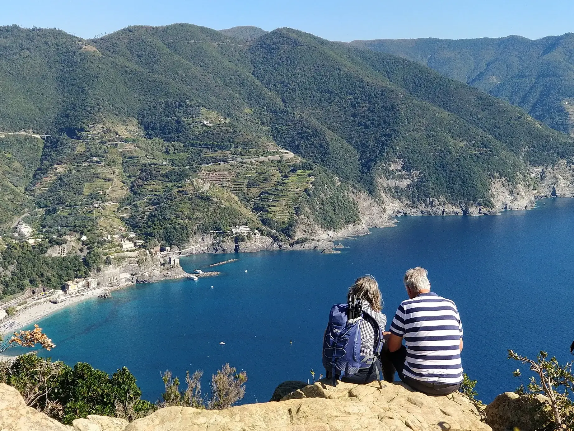 Ein Wanderpaar macht Rast auf einem Felsen und blickt auf die Bucht und Steilküste der Cinque Terre in Ligurien.