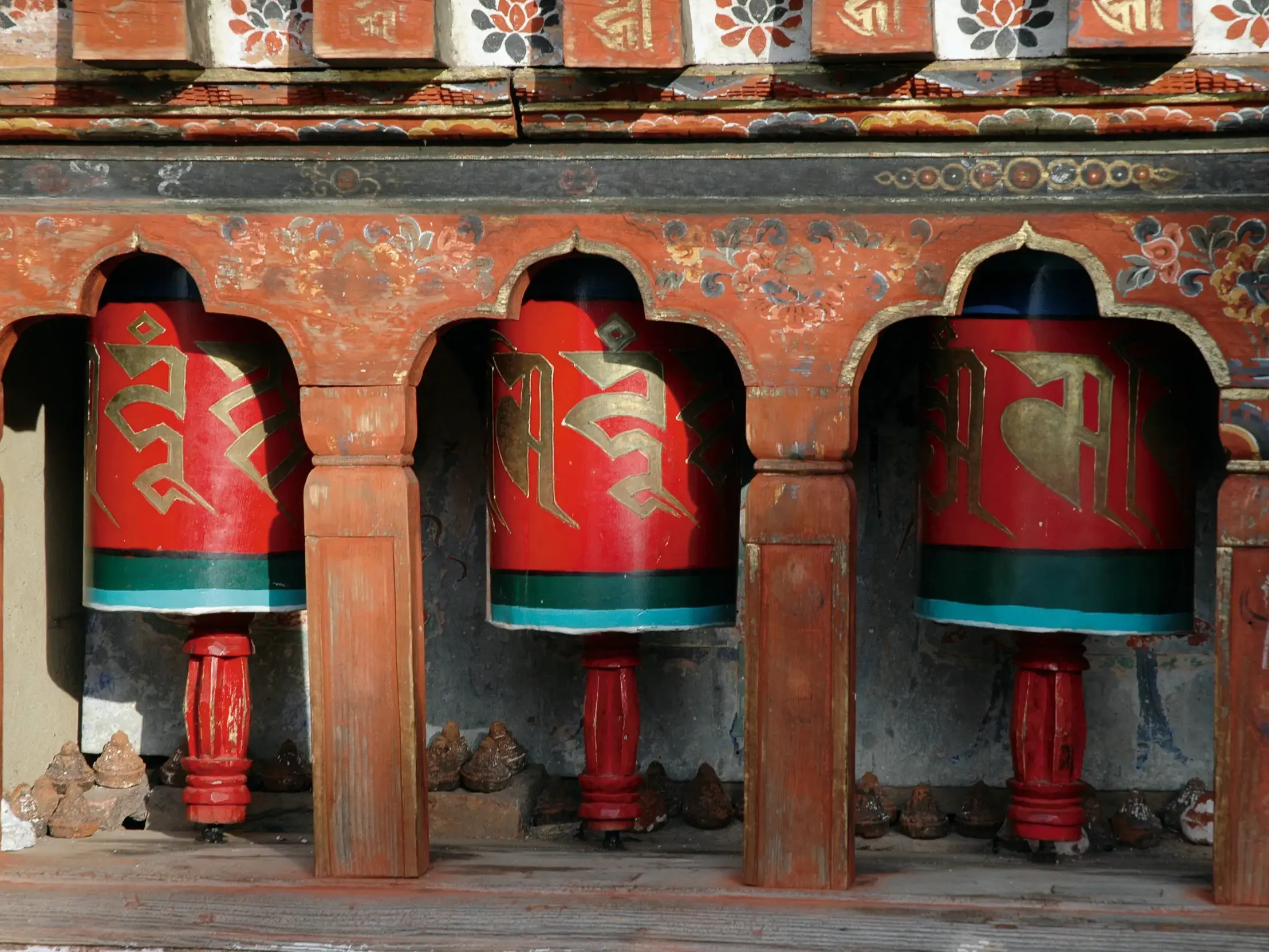 Drei rote Gebetsmühlen mit goldenen Mantras in einem kunstvoll verzierten Holzrahmen in einem Tempel in Bhutan.