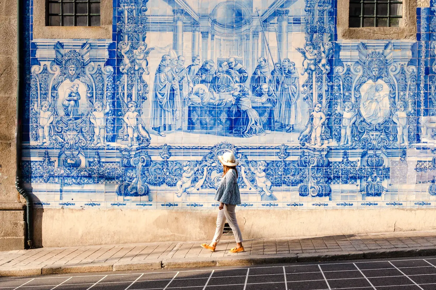 Eine Frau mit Hut spaziert in Porto an einer historischen Wandfassade mit kunstvollen, blau-weißen Azulejo-Fliesen vorbei.