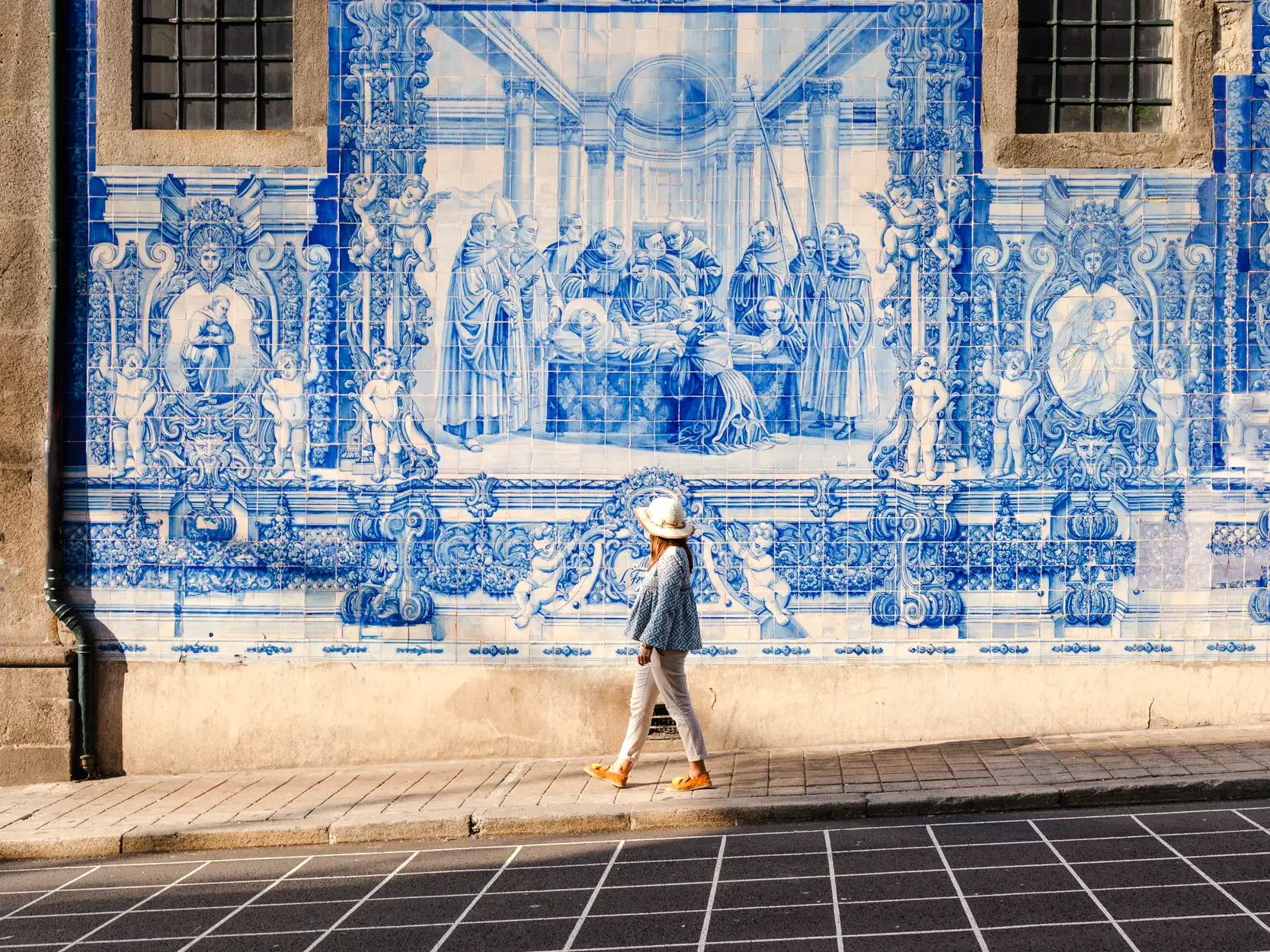 Eine Frau mit Hut spaziert in Porto an einer historischen Wandfassade mit kunstvollen, blau-weißen Azulejo-Fliesen vorbei.