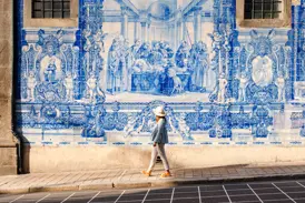 Eine Frau mit Hut spaziert in Porto an einer historischen Wandfassade mit kunstvollen, blau-weißen Azulejo-Fliesen vorbei.