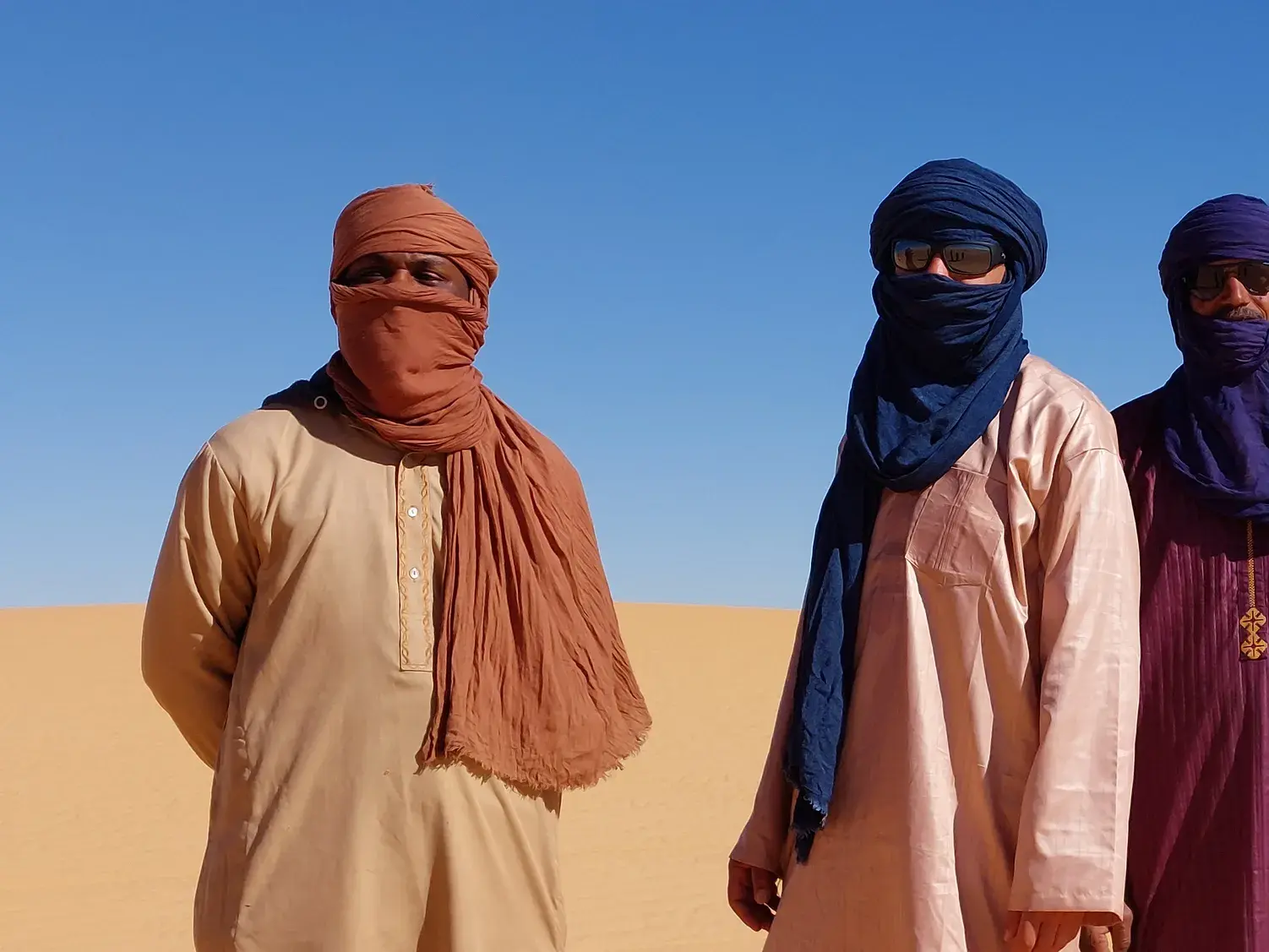Drei Tuareg-Begleiter mit traditionellen Gewändern und Turbanen stehen in der weiten Wüstenlandschaft der Sahara.