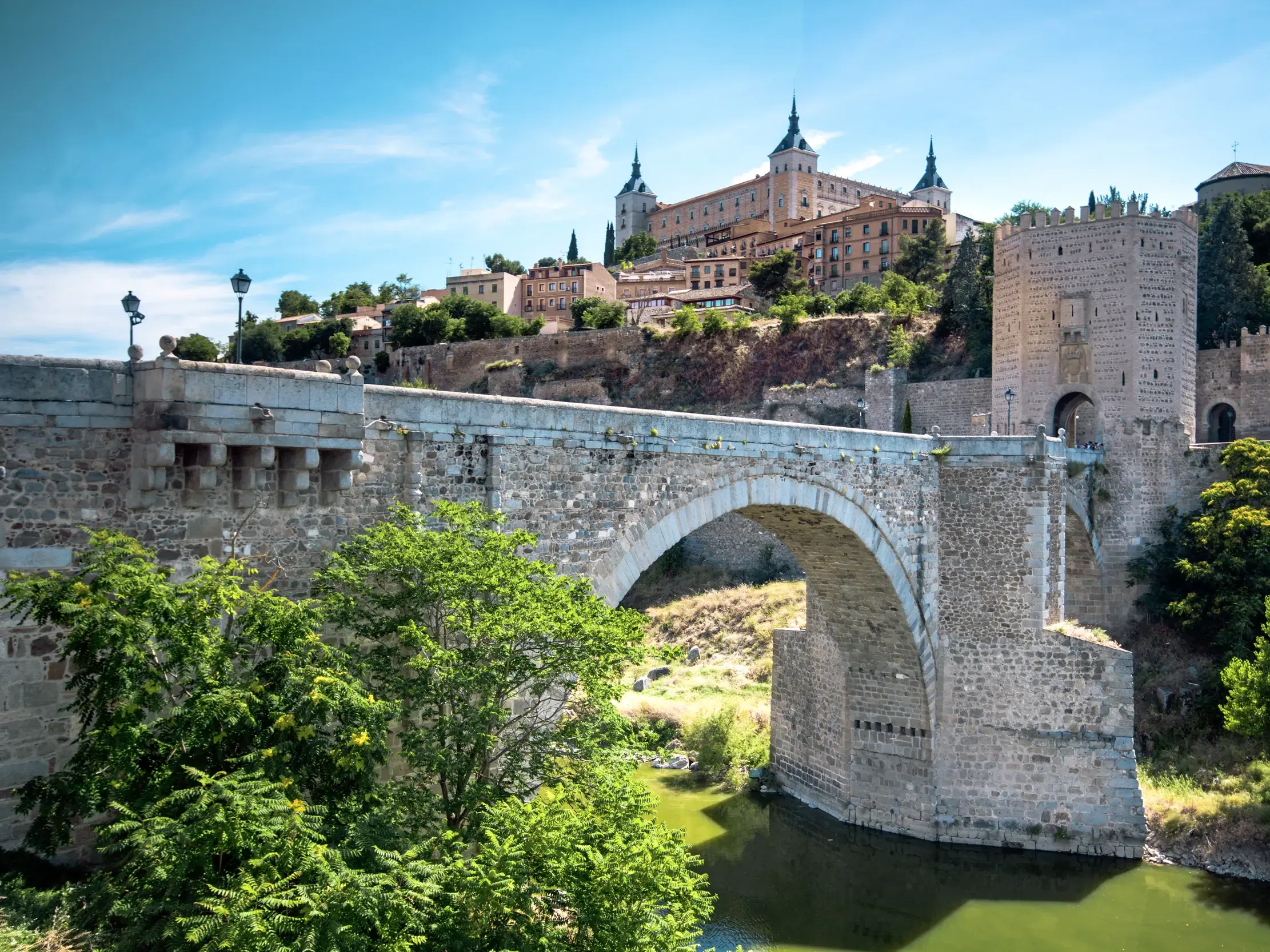Die steinerne Bogenbrücke Puente de San Martín führt über den Tajo zur historischen Altstadt von Toledo unter blauem Himmel.