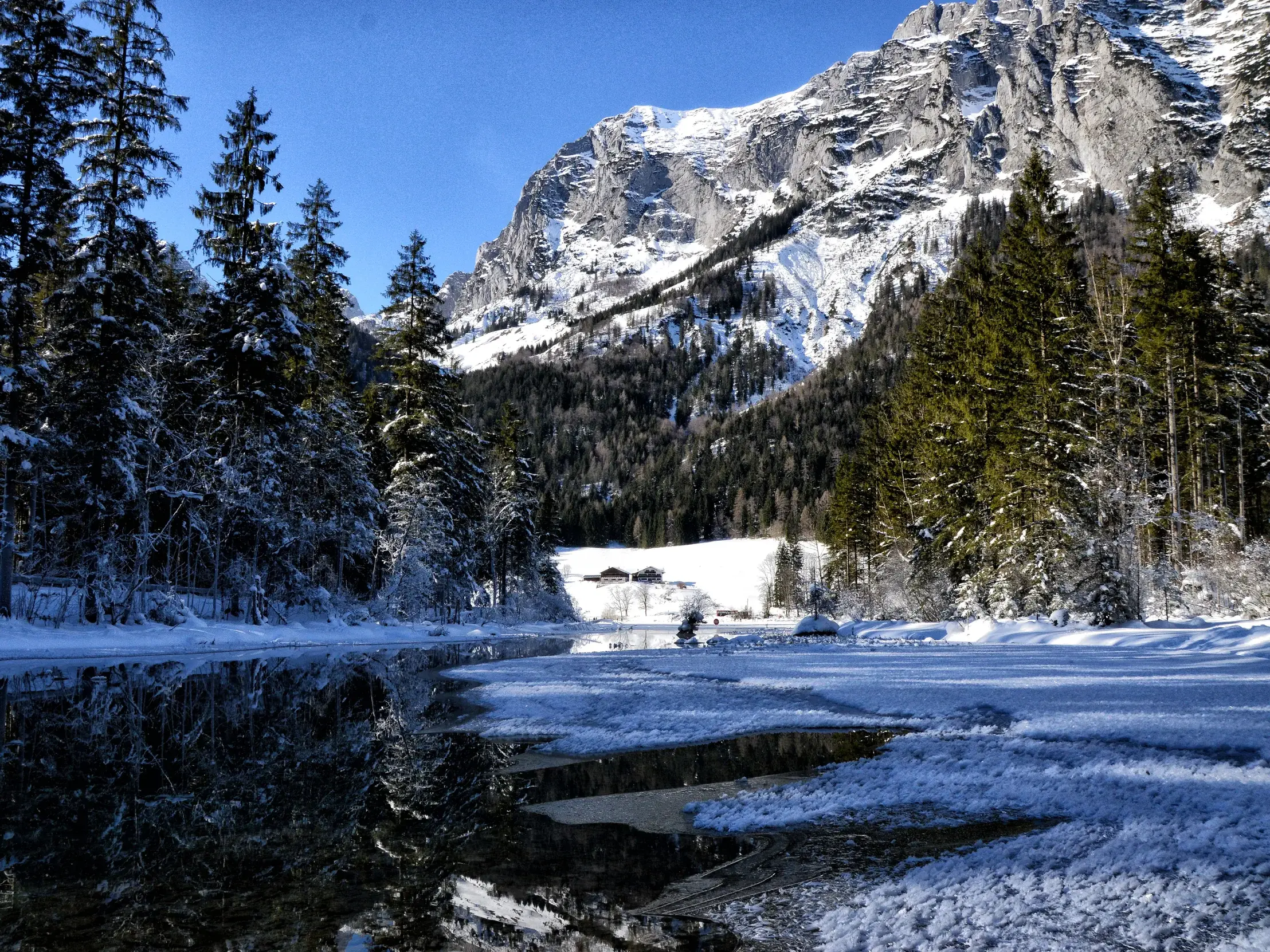 Ein teilweise zugefrorener Fluss spiegelt den verschneiten Wald im Wimbachgries mit sonnigem Blick auf den Hochkalter.