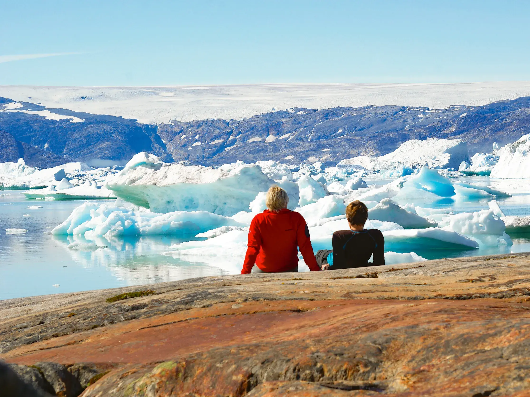 Zwei Wanderer machen eine Pause auf Felsen am Sermilik-Fjord in Ostgrönland und blicken auf Eisberge und das Inlandeis.