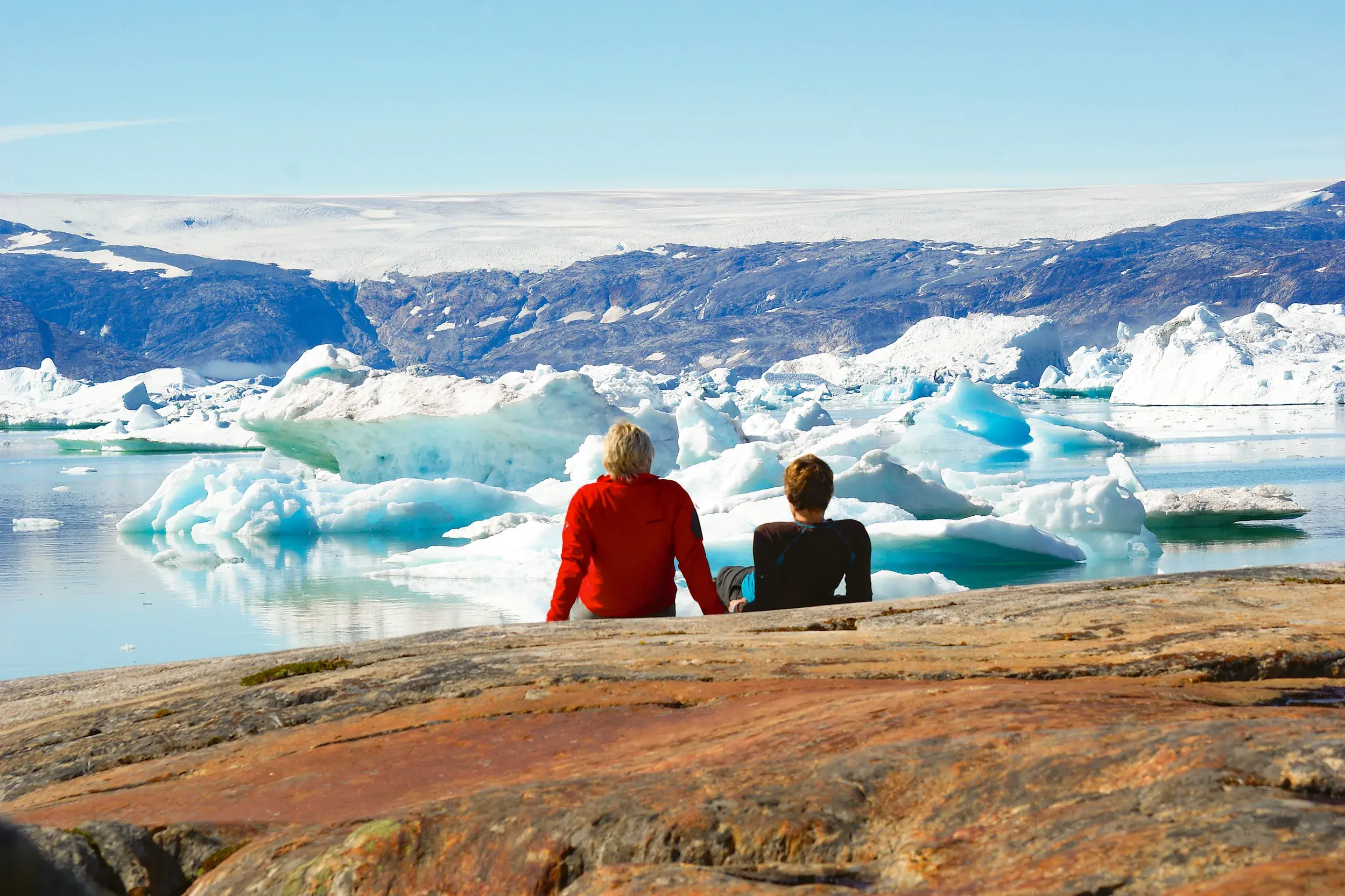 Zwei Wanderer machen eine Pause auf Felsen am Sermilik-Fjord in Ostgrönland und blicken auf Eisberge und das Inlandeis.