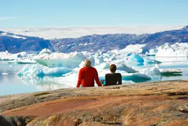 Zwei Wanderer machen eine Pause auf Felsen am Sermilik-Fjord in Ostgrönland und blicken auf Eisberge und das Inlandeis.