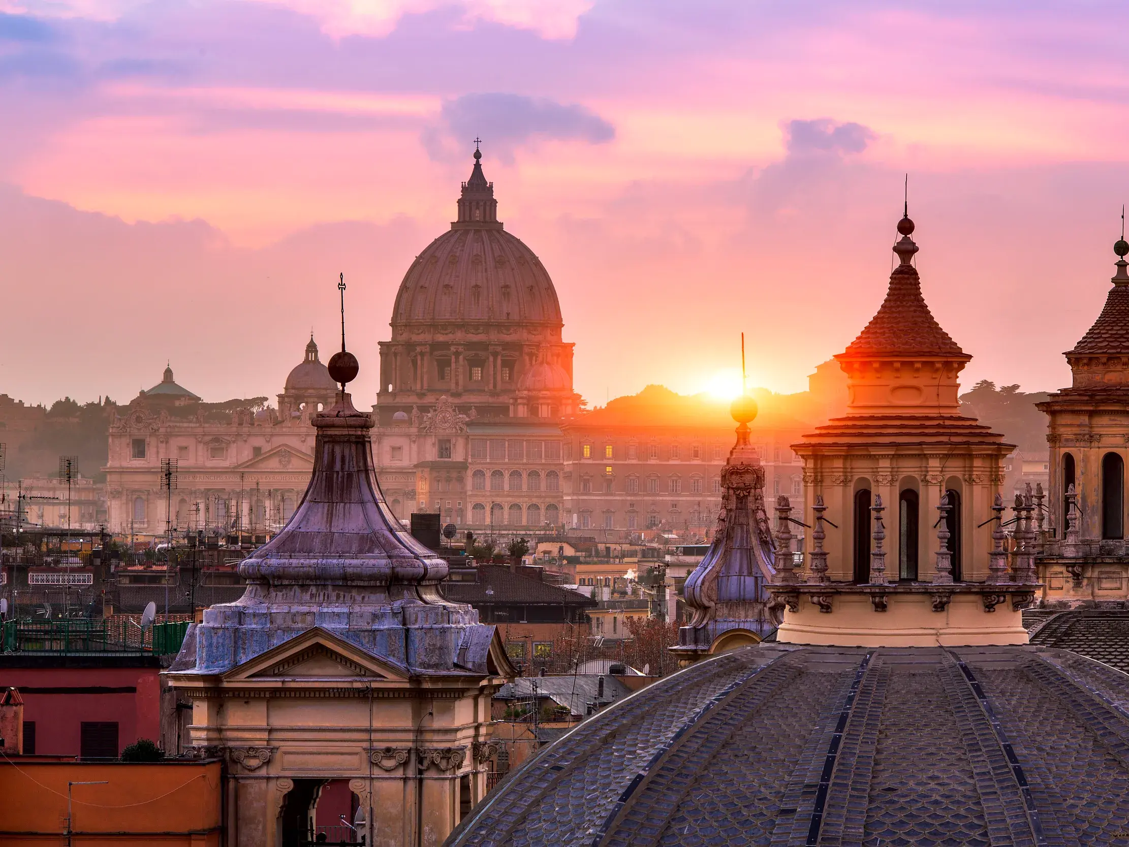 Malerischer Sonnenuntergang über Rom mit Blick auf die Kuppel der Peterskirche und weitere Türme und Kuppeln im Vordergrund.