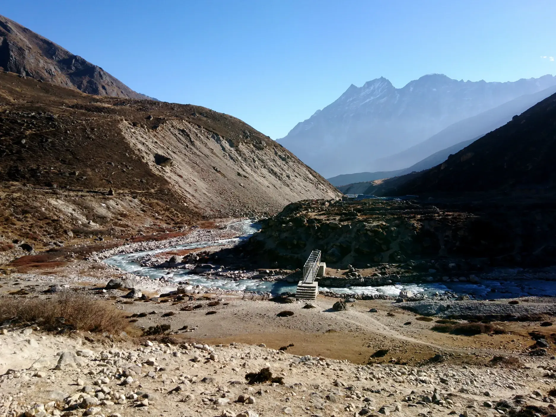Trekkingpfad in Nepal führt über eine Brücke im Bhote Koshi-Tal, umgeben von kargen Hängen und hohen Himalaya-Gipfeln.