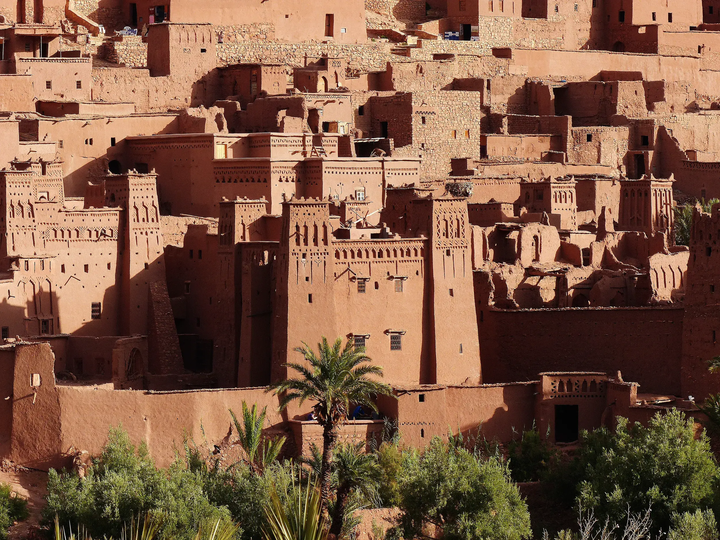 Die historischen Lehmbauten der Kasbah Ait Benhaddou in Marokko thronen bei warmem Sonnenlicht über einer grünen Palmenoase.