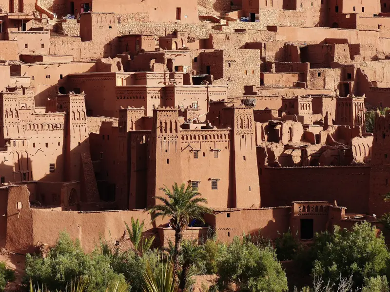 Die historischen Lehmbauten der Kasbah Ait Benhaddou in Marokko thronen bei warmem Sonnenlicht über einer grünen Palmenoase.