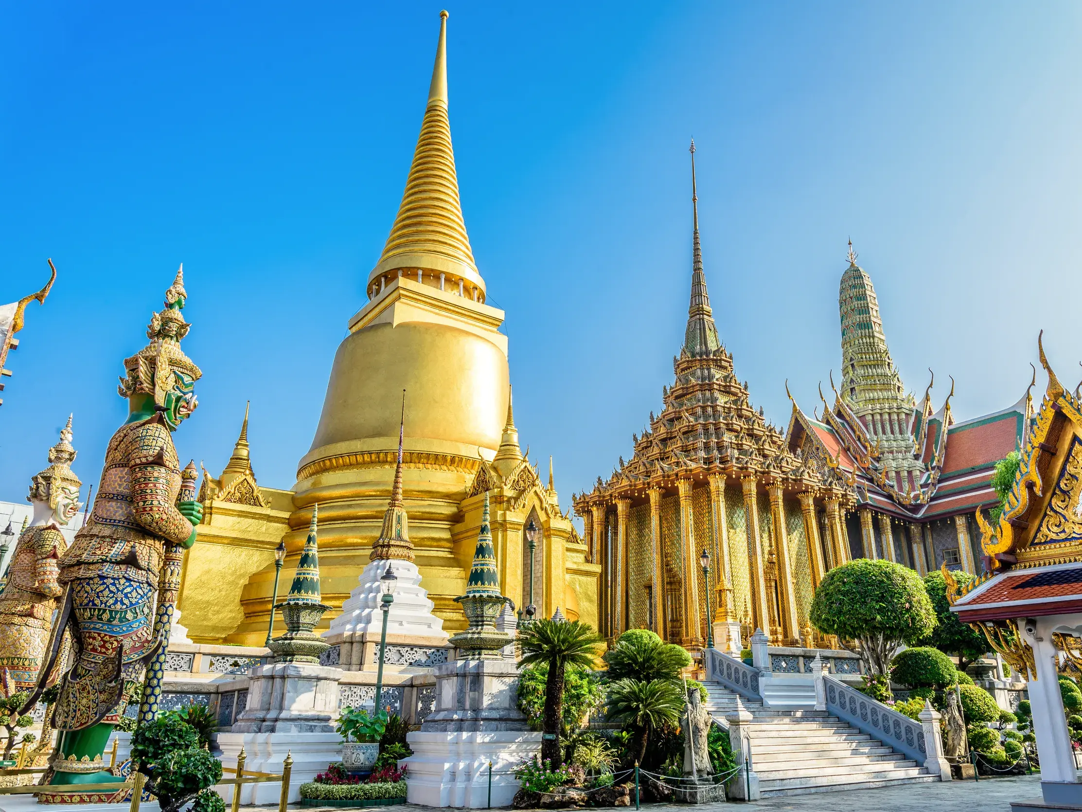 Prachtvoller goldener Chedi und Tempel im Grand Palace in Bangkok, bewacht von einer Yaksha-Statue.
