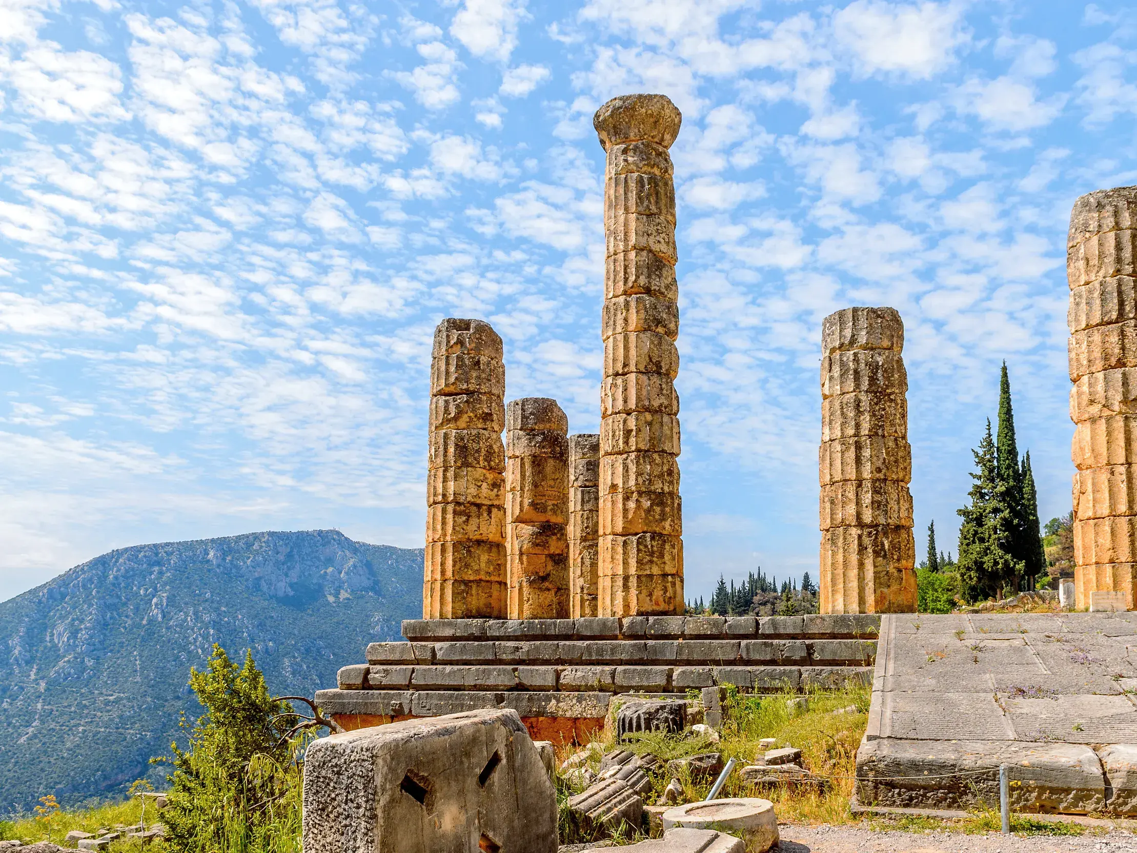 Antike Säulen der Orakelstätte Delphi thronen auf einem Hügel vor einer Bergkulisse unter leicht bewölktem Himmel.
