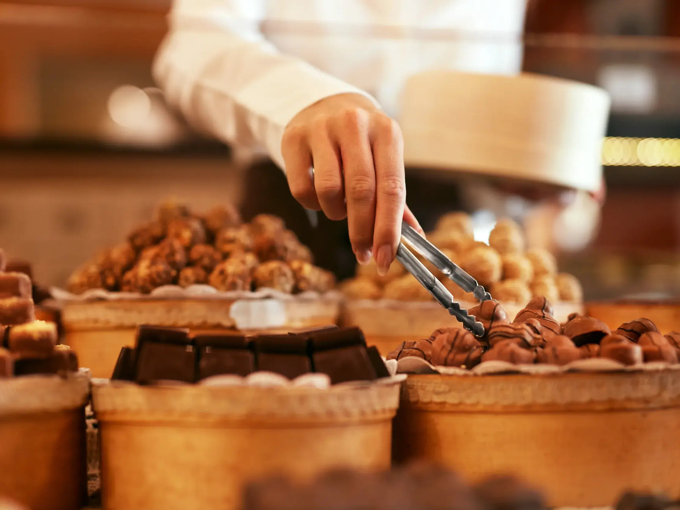 Eine Chocolatière wählt mit einer Zange eine feine Praline aus der Theke einer belgischen Schokoladen-Manufaktur.