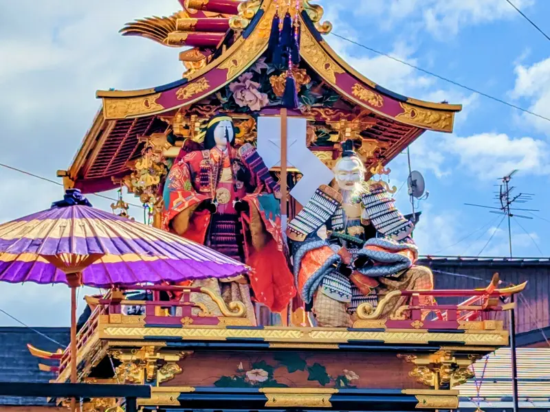 Kunstvoll verzierter Festwagen in Takayama mit traditionellen Figuren in Kimono und Samurai-Rüstung vor blauem Himmel.