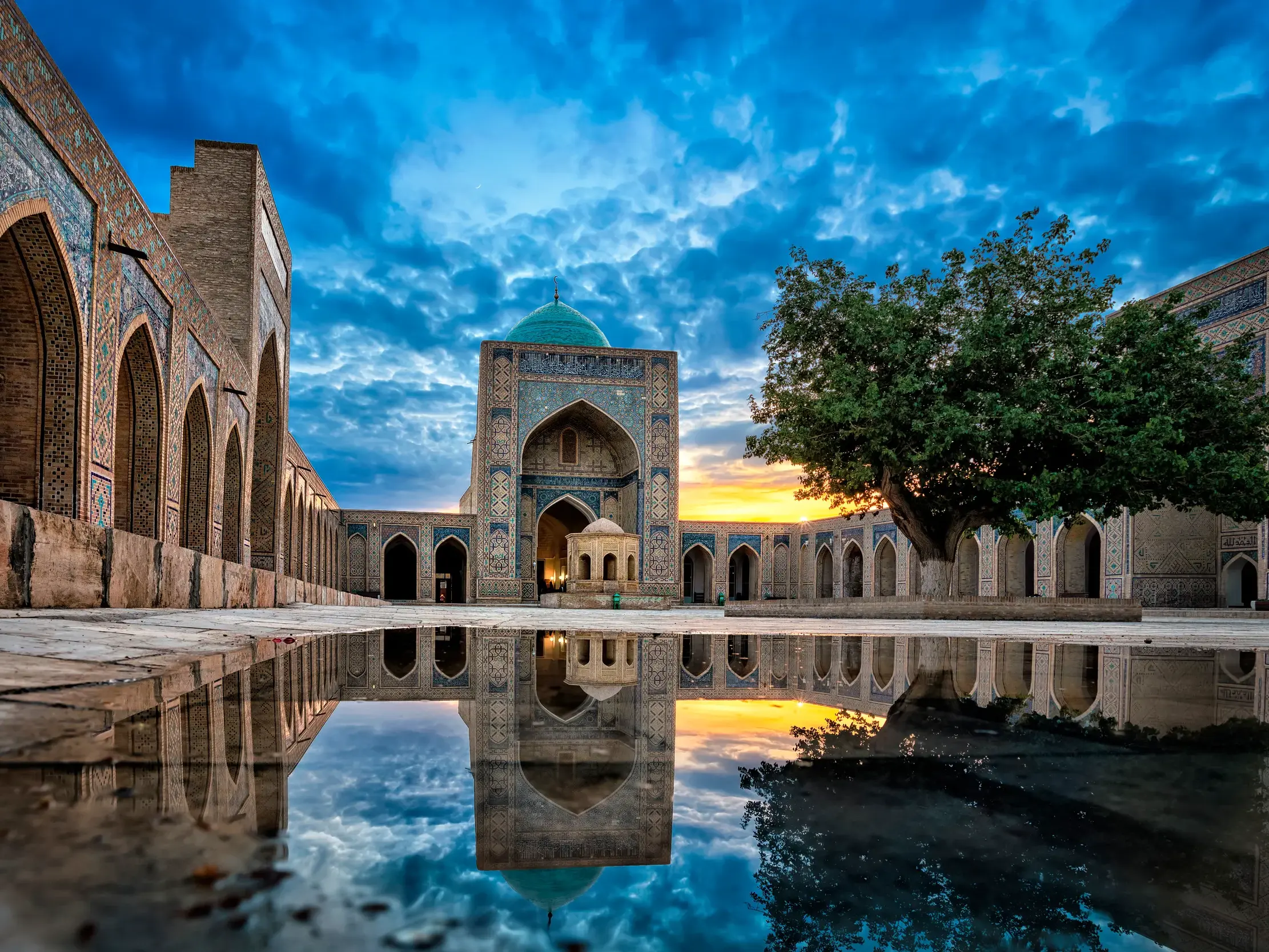 Historische Moschee mit türkiser Kuppel in Buchara, die sich bei Sonnenuntergang in einer Wasserpfütze auf dem Vorplatz spiegelt.