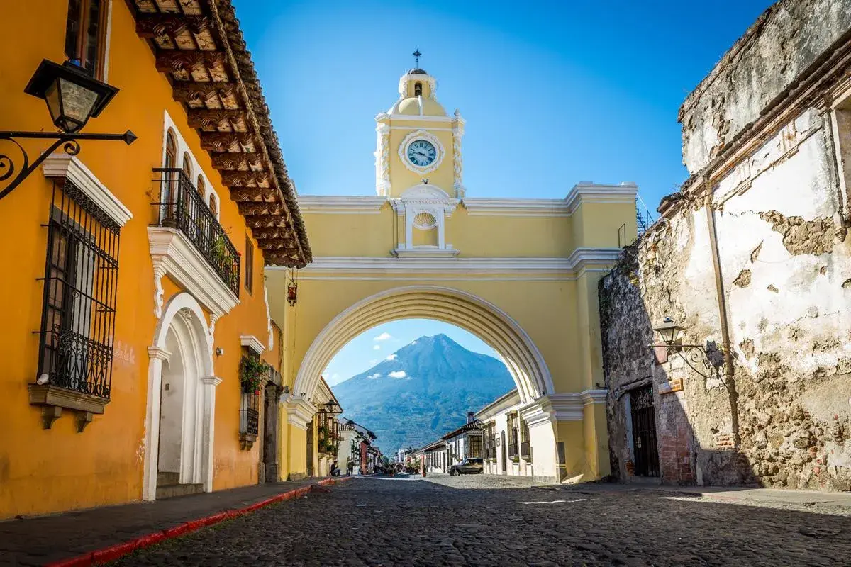 Kopfsteinpflasterstraße in Antigua mit dem gelben Arco de Santa Catalina, der den Vulkan Agua an einem sonnigen Tag einrahmt.