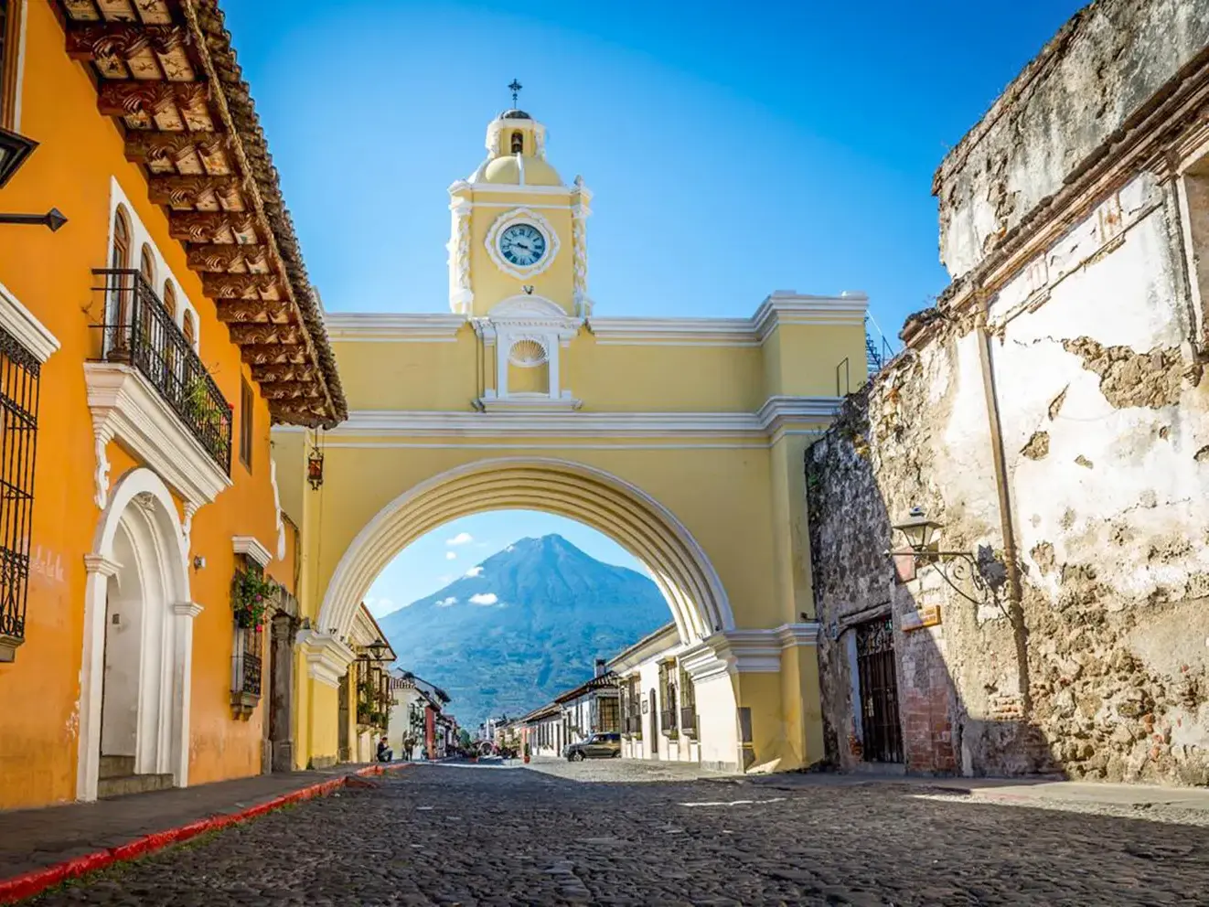 Kopfsteinpflasterstraße in Antigua mit dem gelben Arco de Santa Catalina, der den Vulkan Agua an einem sonnigen Tag einrahmt.