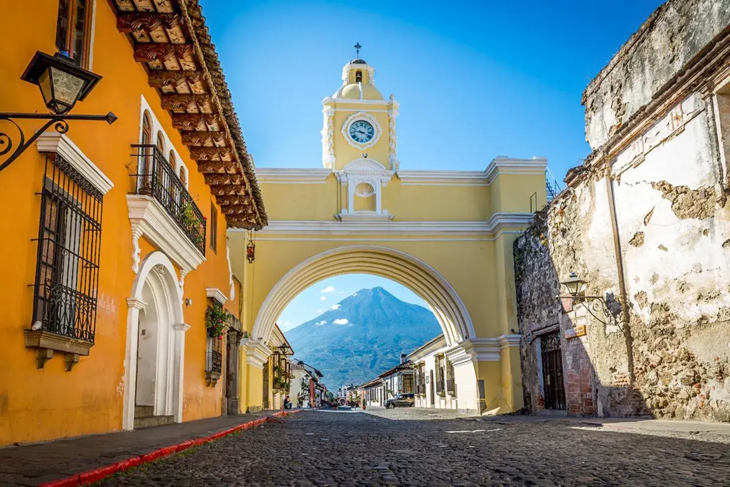 Kopfsteinpflasterstraße in Antigua mit dem gelben Arco de Santa Catalina, der den Vulkan Agua an einem sonnigen Tag einrahmt.