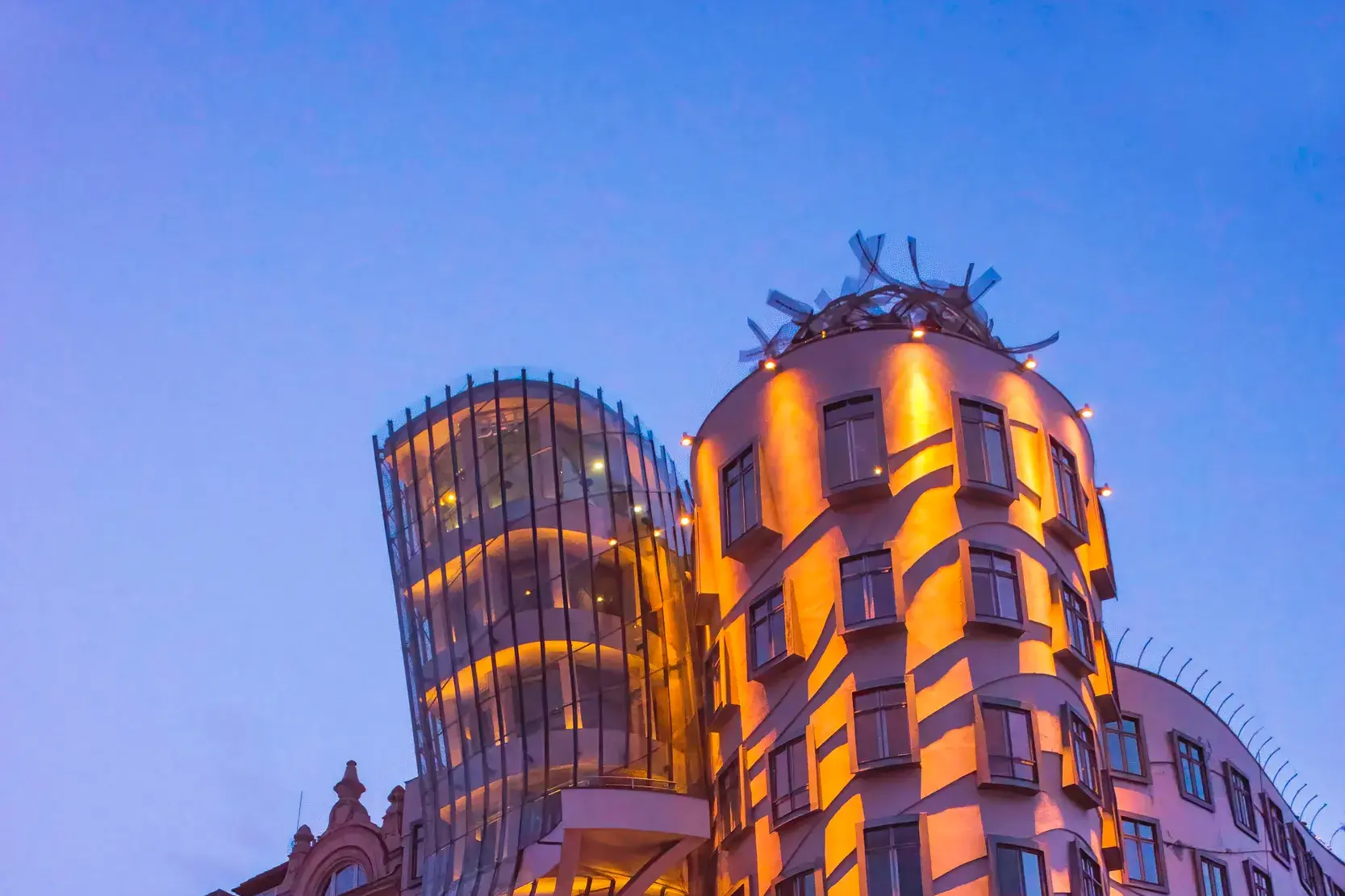 Das moderne Tanzende Haus in Prag leuchtet bei Dämmerung in warmen Farben neben einem klassischen Altbau an der Moldau.