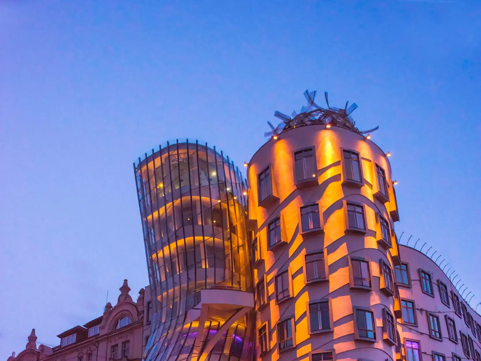 Das moderne Tanzende Haus in Prag leuchtet bei Dämmerung in warmen Farben neben einem klassischen Altbau an der Moldau.