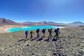 Sechs Bergsteiger einer Trekking-Gruppe wandern mit Stöcken an der türkisfarbenen Laguna Verde in der Atacama-Wüste in Chile.