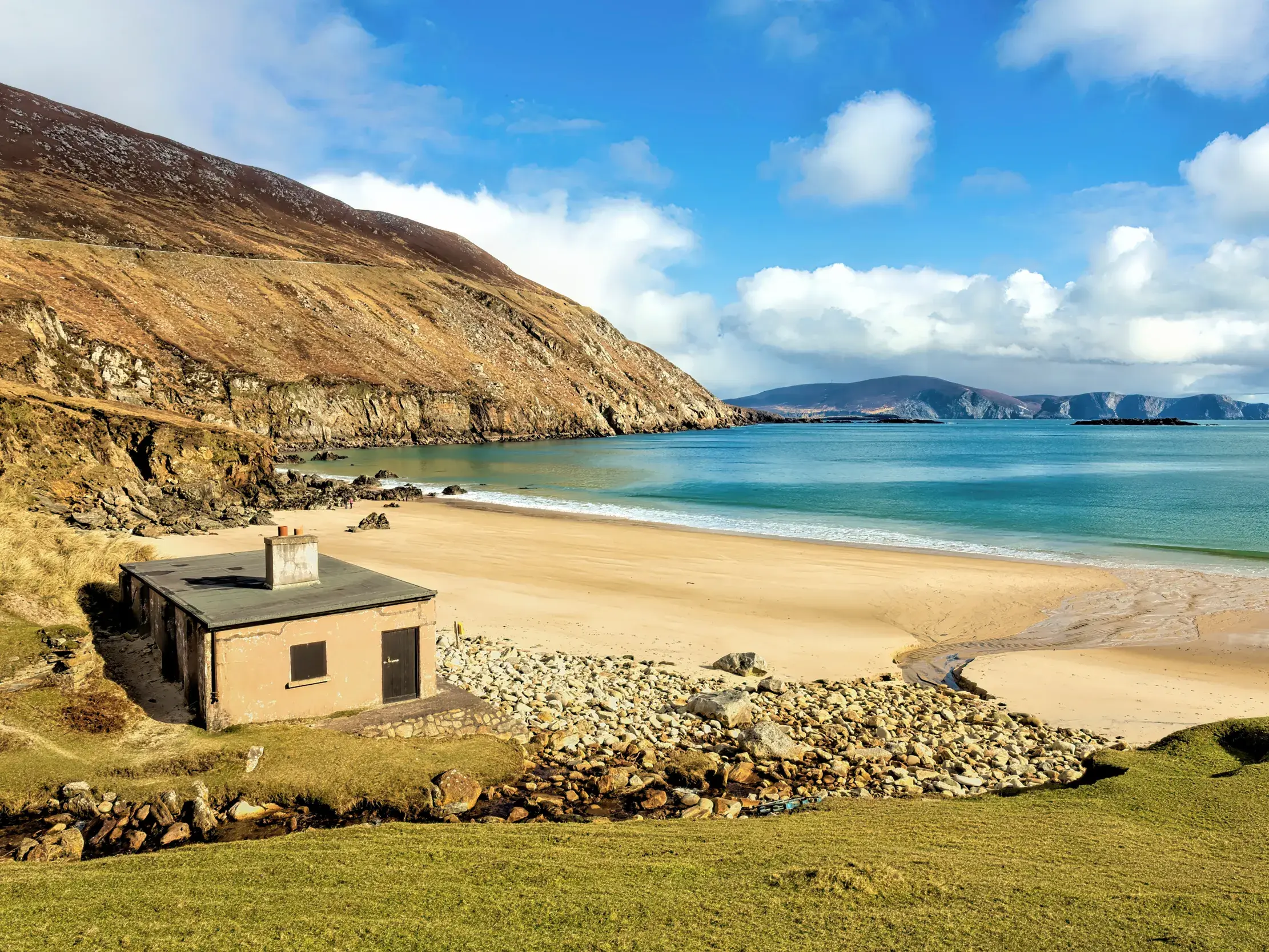 Malerische Bucht Keem Beach auf Achill Island in Irland mit einem kleinen Haus am Sandstrand vor rauen, grünen Klippen.