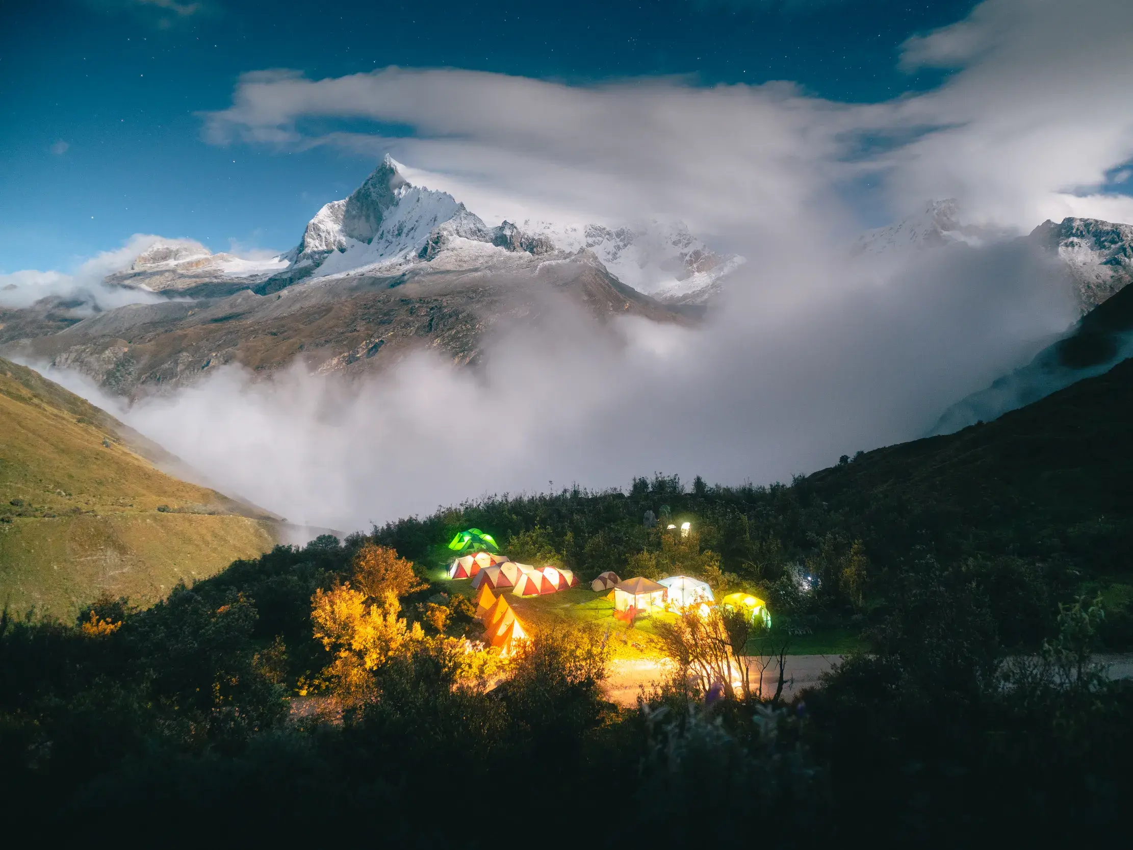 Ein beleuchtetes Camp mit Luxuszelten in der peruanischen Cordillera Blanca bei Nacht vor der Kulisse eines schneebedeckten Gipfels.