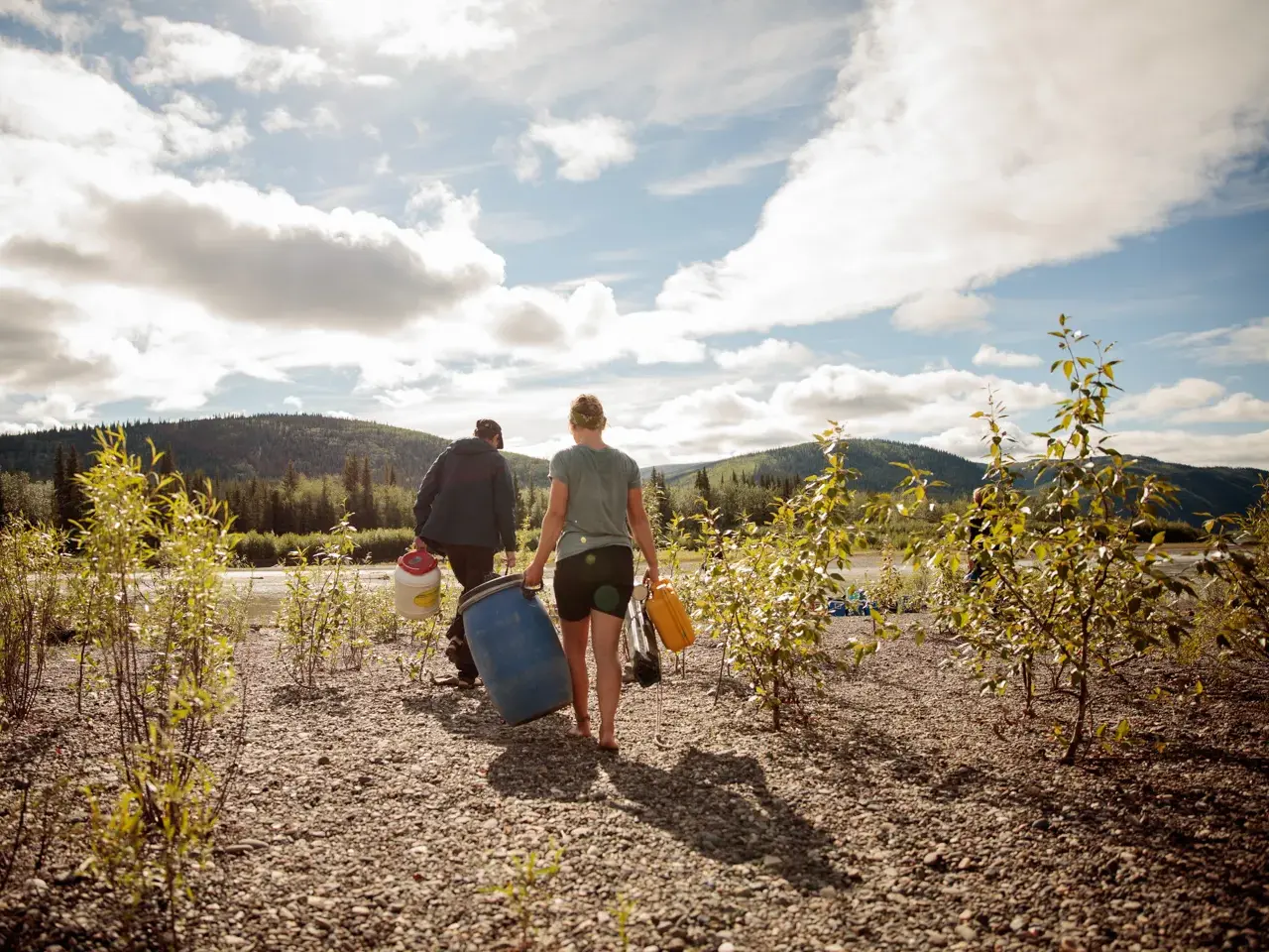Eine Frau und ein Mann tragen wasserdichte Tonnen und Packsäcke zum Startpunkt des Kanutrekkings am Yukon River.