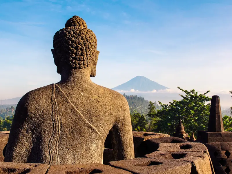 Blick von der buddhistischen Tempelanlage BOROBUDUR über eine Buddha-Statue hinweg auf einen Vulkan im Morgennebel auf Java.