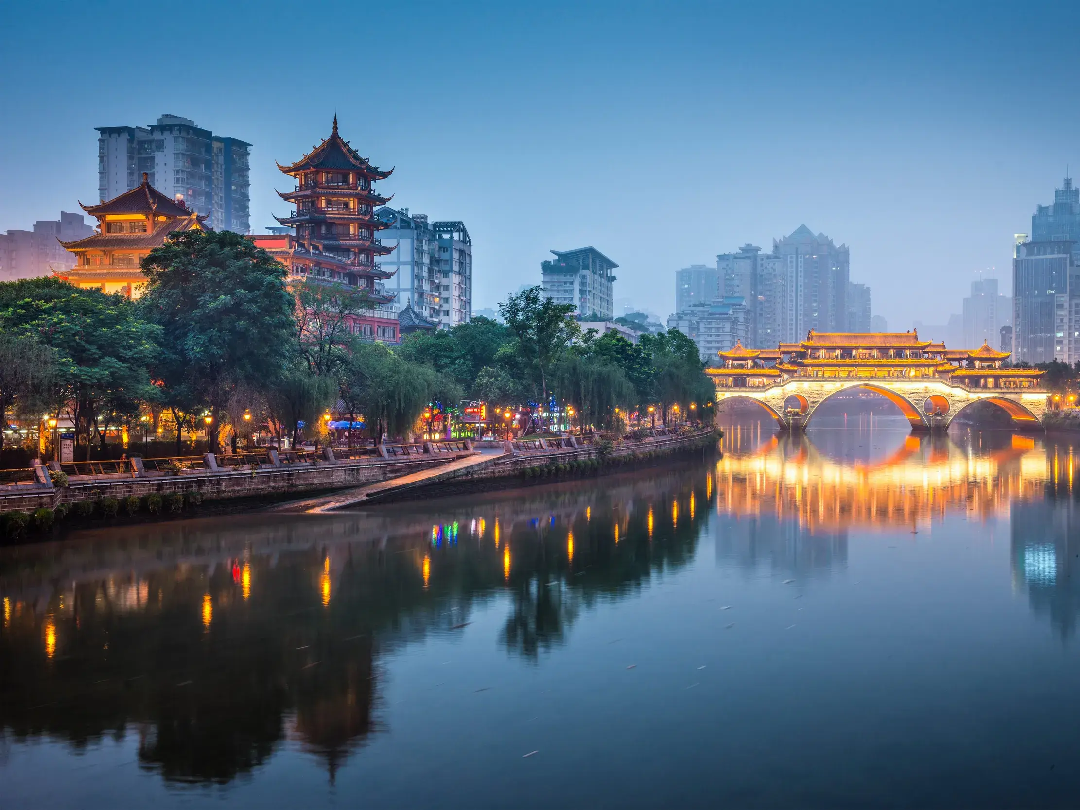 Die historische Anshun-Brücke in Chengdu spannt sich beleuchtet über den Jin-Fluss bei Dämmerung, im Hintergrund die Skyline.