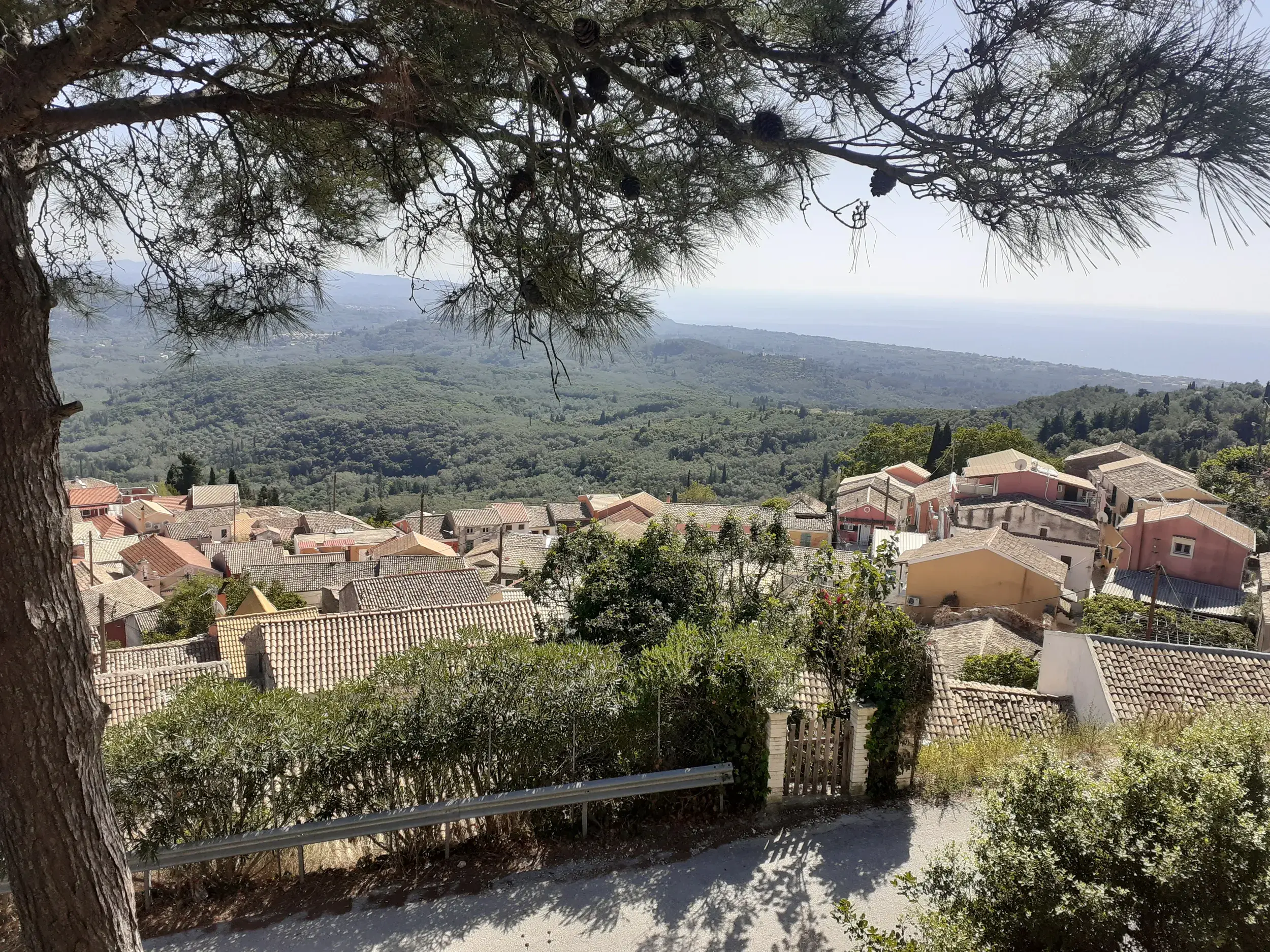 Beim Wandern auf dem Korfu-Trail: Panoramablick über ein kleines Dorf, grüne Hügel und das Ionische Meer in der Ferne.