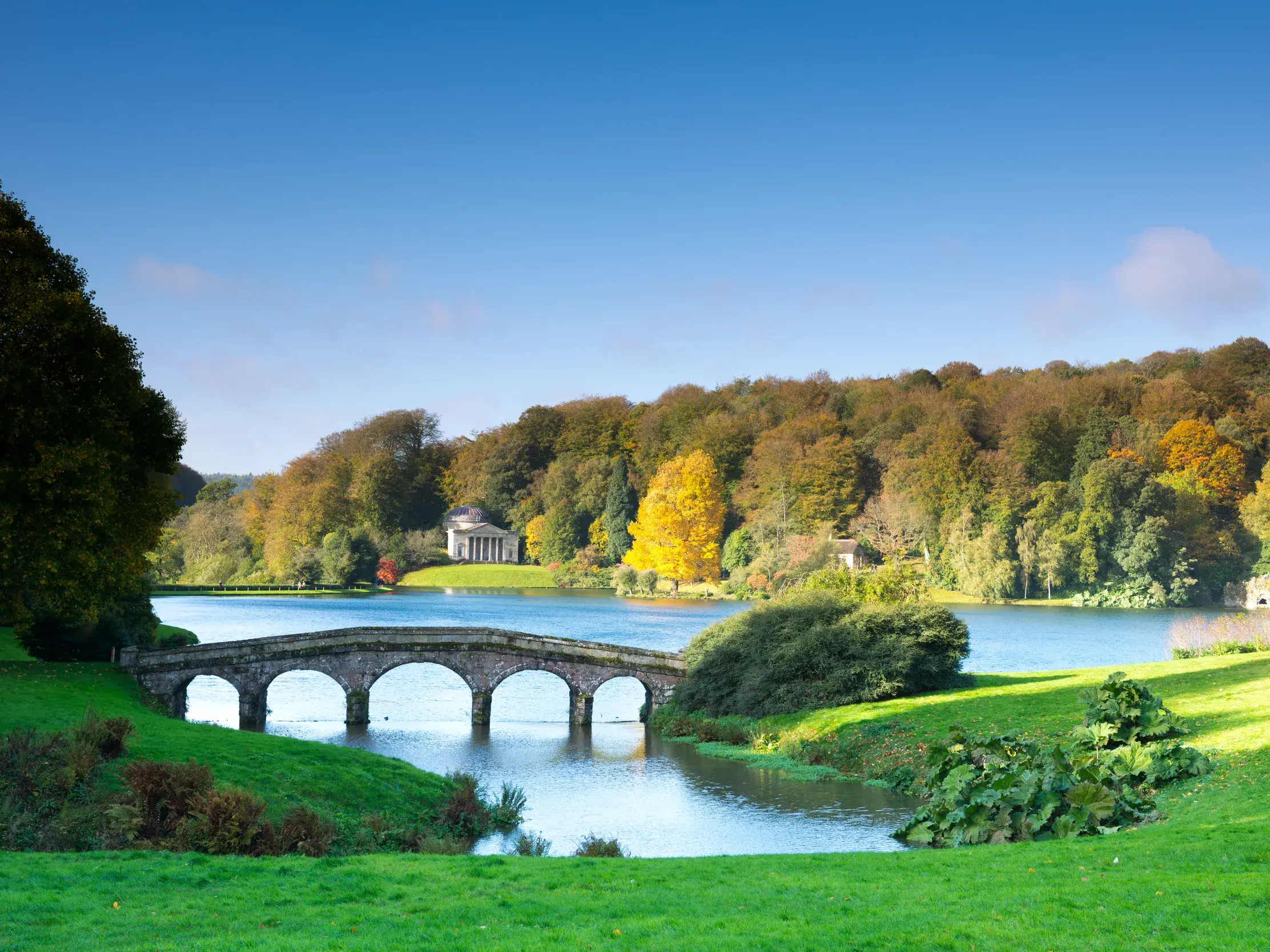 Malerischer Landschaftsgarten von Stourhead in Südengland mit der Steinbrücke und dem Tempel des Apollo am Seeufer.