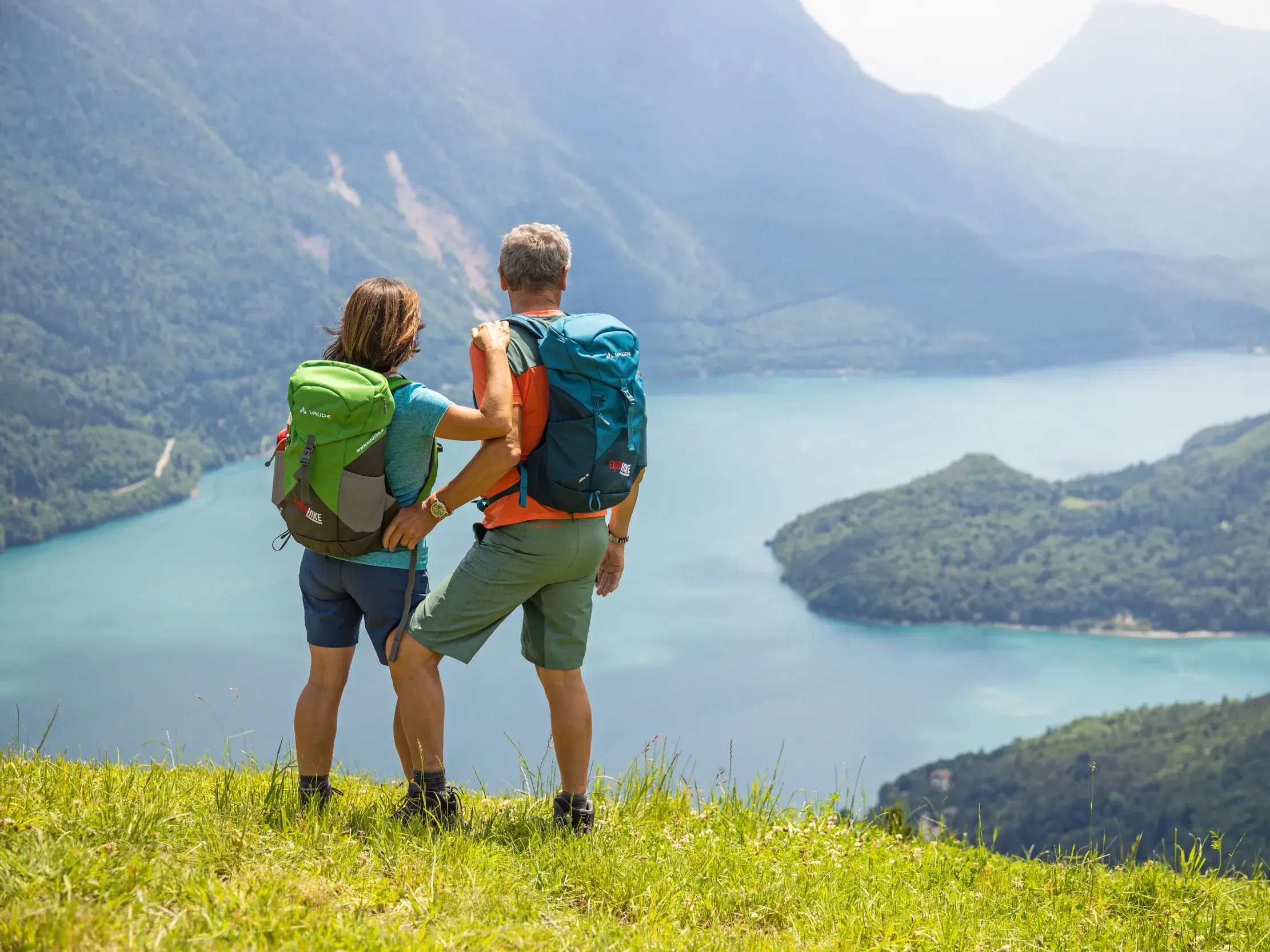 Ein Wander-Paar mit Rucksäcken blickt von einer Bergwiese auf den türkisblauen Gardasee im Trentino.