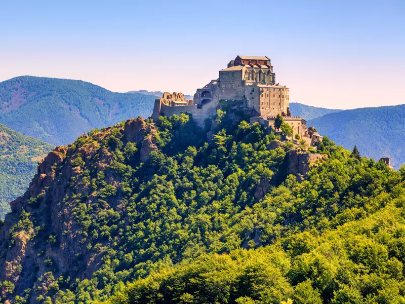 Die Klosteranlage Sacra di San Michele thront bei Sonnenschein auf einem Felsgipfel über dem grünen Susatal im Piemont.