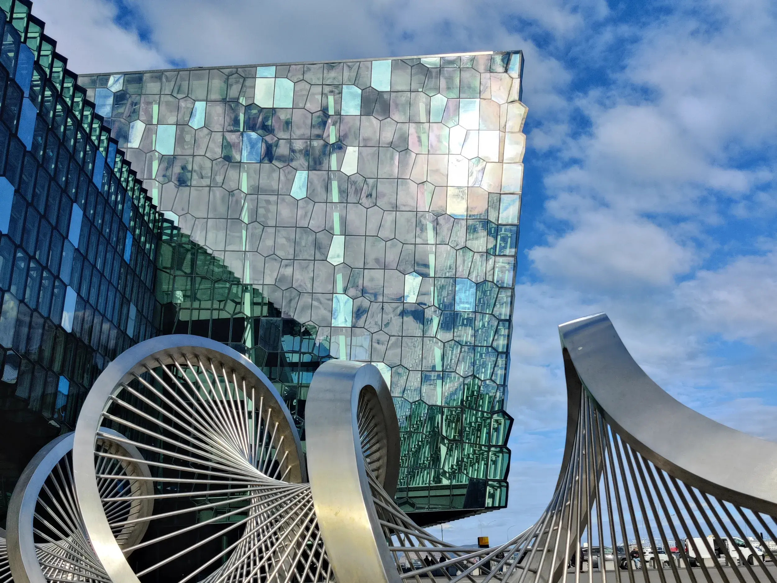 Moderne Skulptur vor der markanten Glasfassade des Konzerthauses Harpa in Reykjavik unter einem strahlend blauen Himmel.