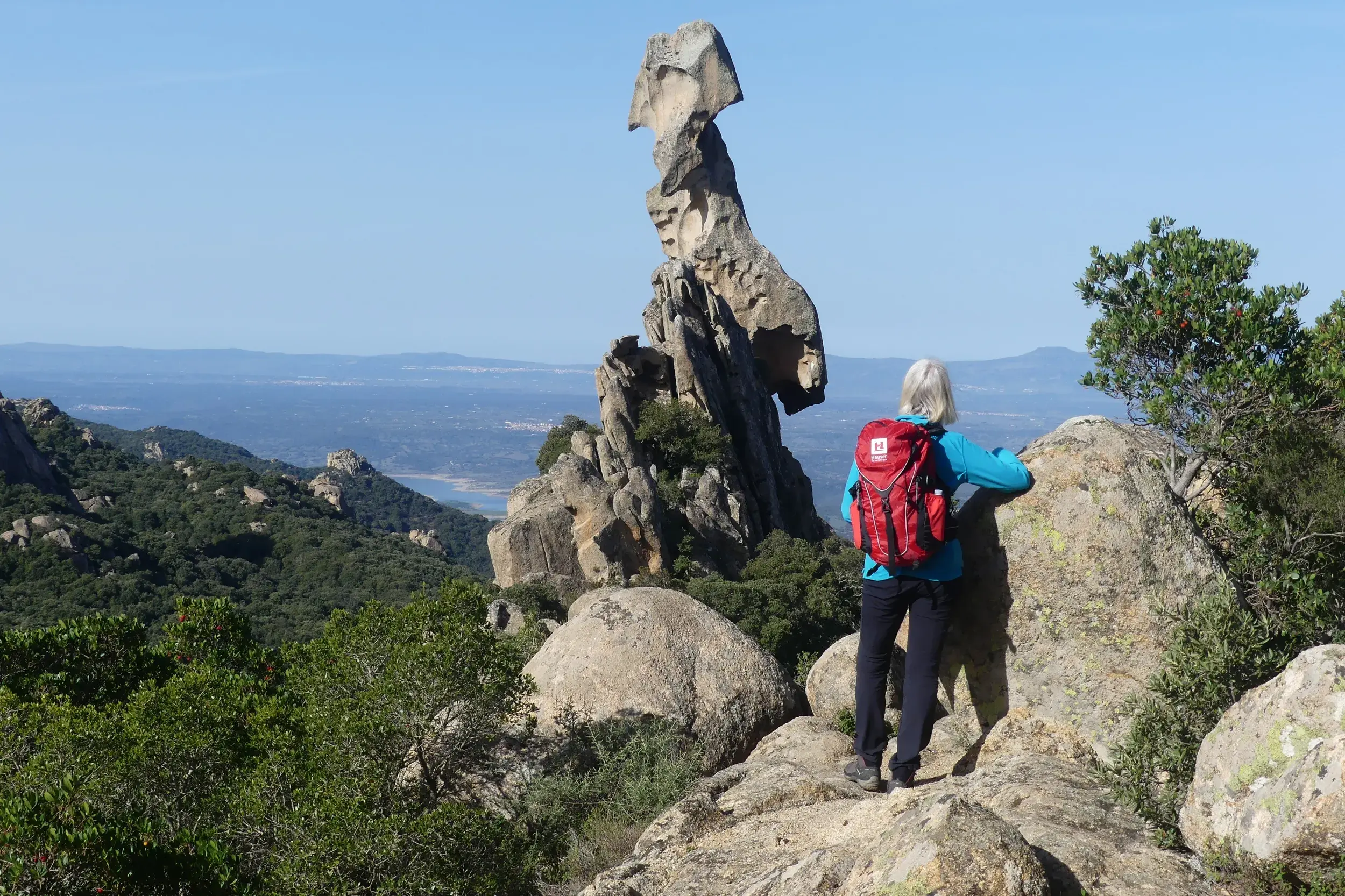 Wanderin mit rotem Rucksack blickt auf den Monolith Sa Carbrarissa in den Wäldern von Austis auf Sardinien.