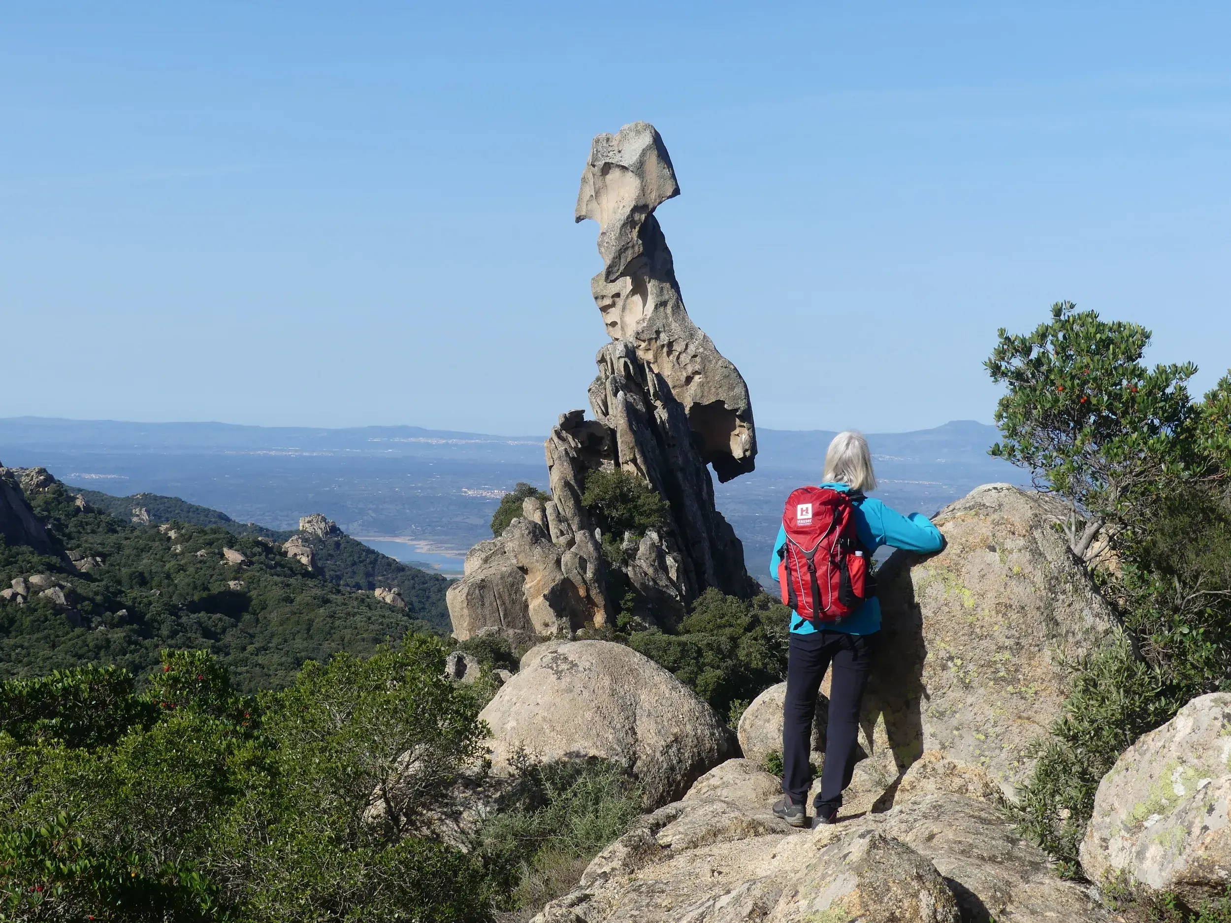 Wanderin mit rotem Rucksack blickt auf den Monolith Sa Carbrarissa in den Wäldern von Austis auf Sardinien.