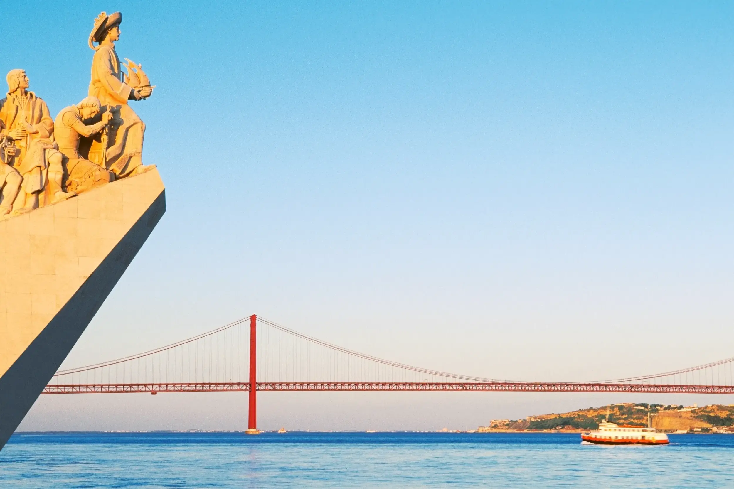 Sonnige Ansicht des Denkmals der Entdeckungen am Tejo in Lissabon mit der ikonischen Tejo-Brücke und einem Boot im Hintergrund.