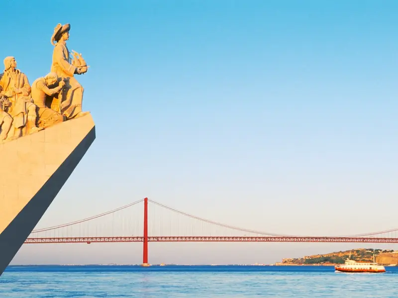Sonnige Ansicht des Denkmals der Entdeckungen am Tejo in Lissabon mit der ikonischen Tejo-Brücke und einem Boot im Hintergrund.